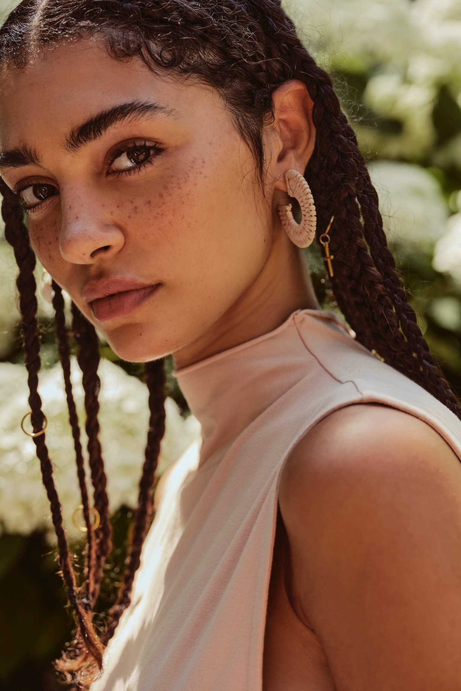 A woman with dark curly hair and freckles looking at the camera, wearing a sleeveless light-colored top and unique hoop earrings, outdoors with blurred green foliage in the background.