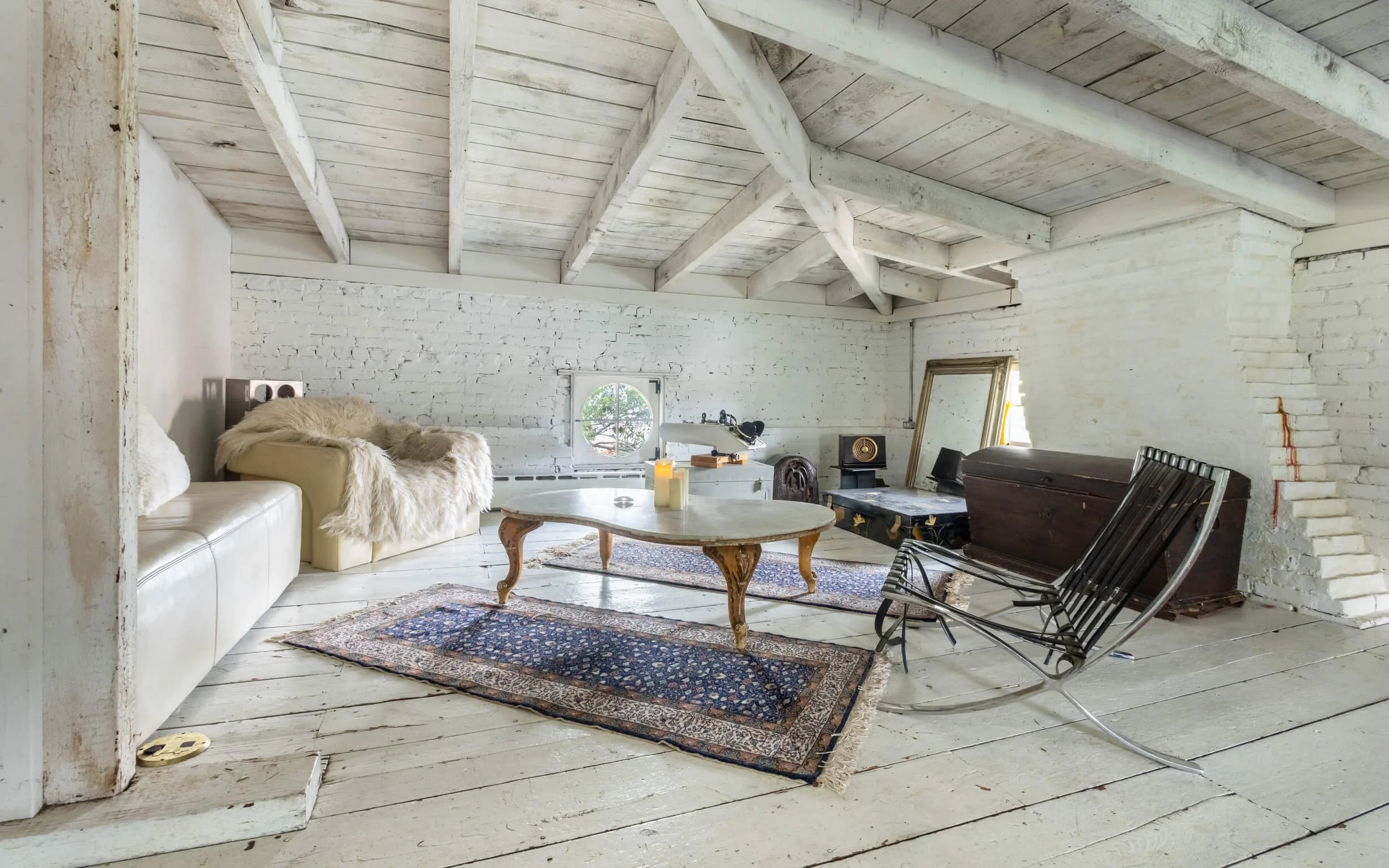 Living room with white brick walls and vaulted white wooden ceiling, featuring a white sofa with a furry throw, an ornate coffee table, a black rocking chair, a vintage chest, a large mirror, a window, and various vintage and modern decor items.