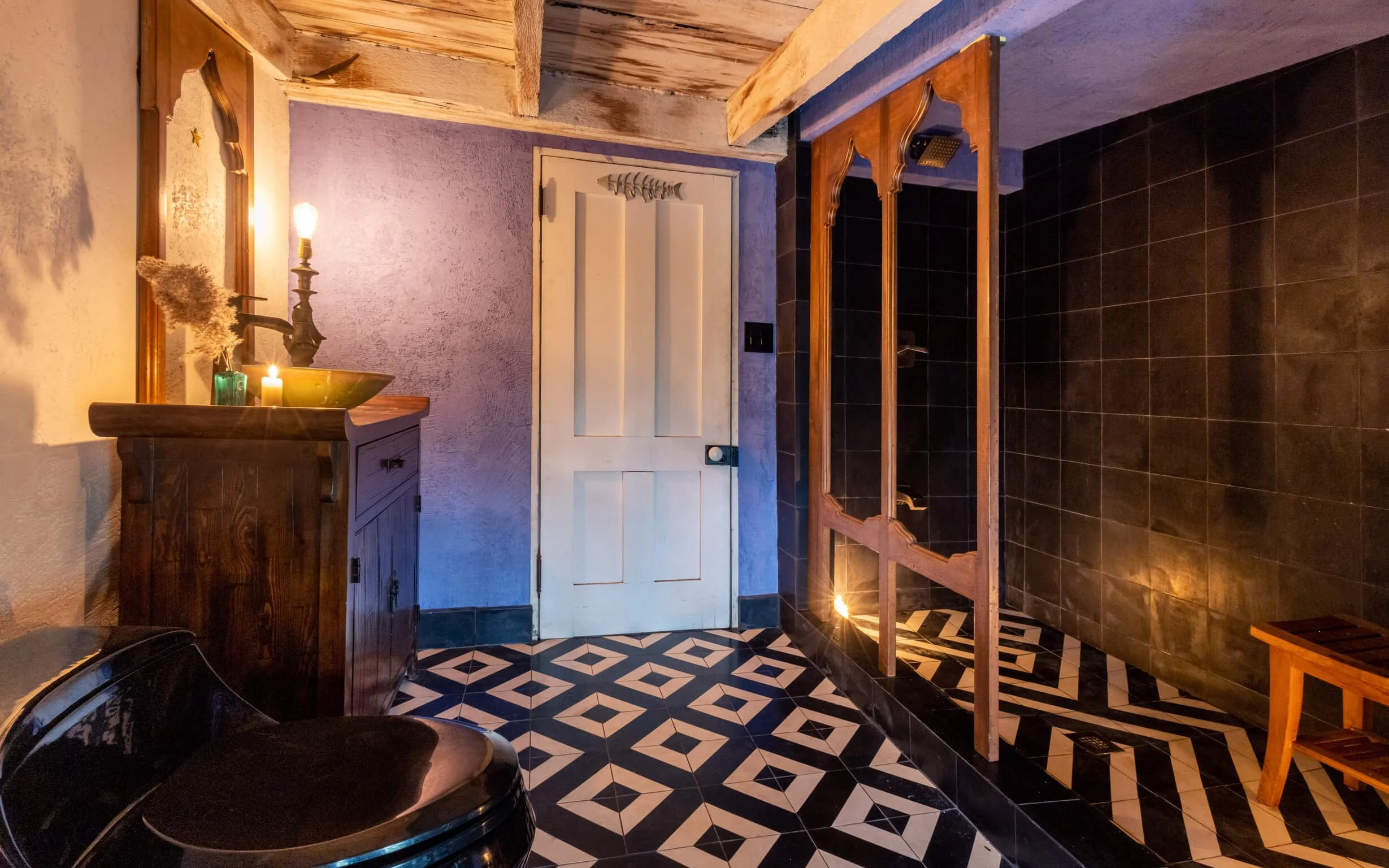 A rustic bathroom with patterned black and white tiled floor, purple textured wall, and wooden accents including a vanity, mirror, and shower enclosure. Candles and decorative items are on the vanity.