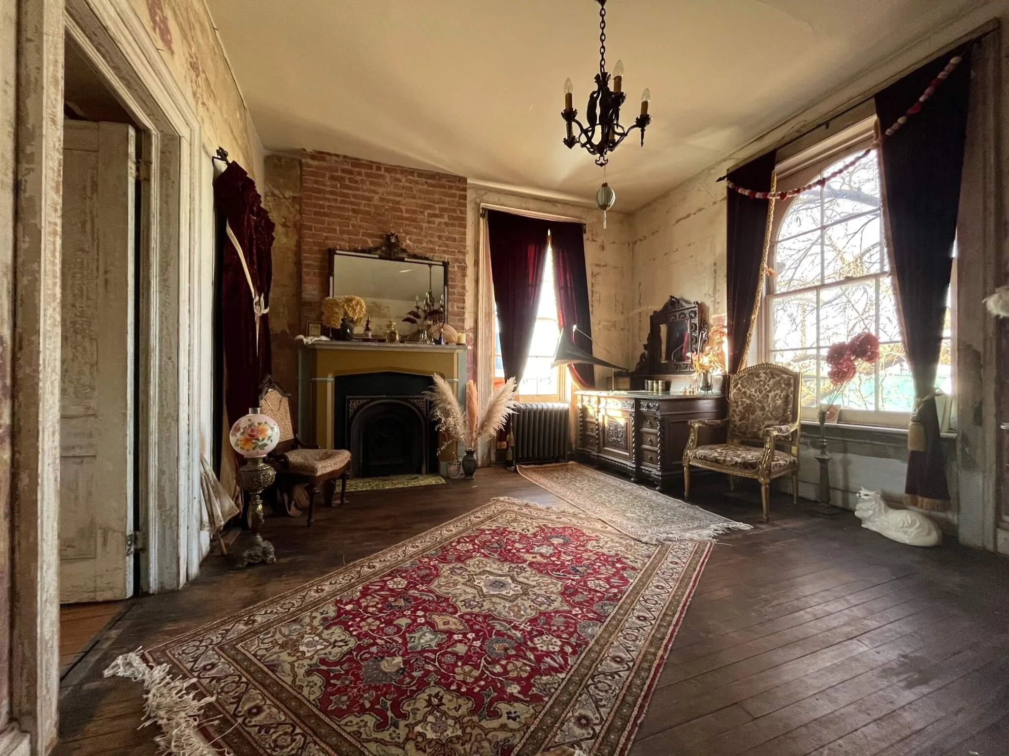 Vintage living room with antique furniture, ornate rug, fireplace, large windows with dark curtains, and decorative plants.