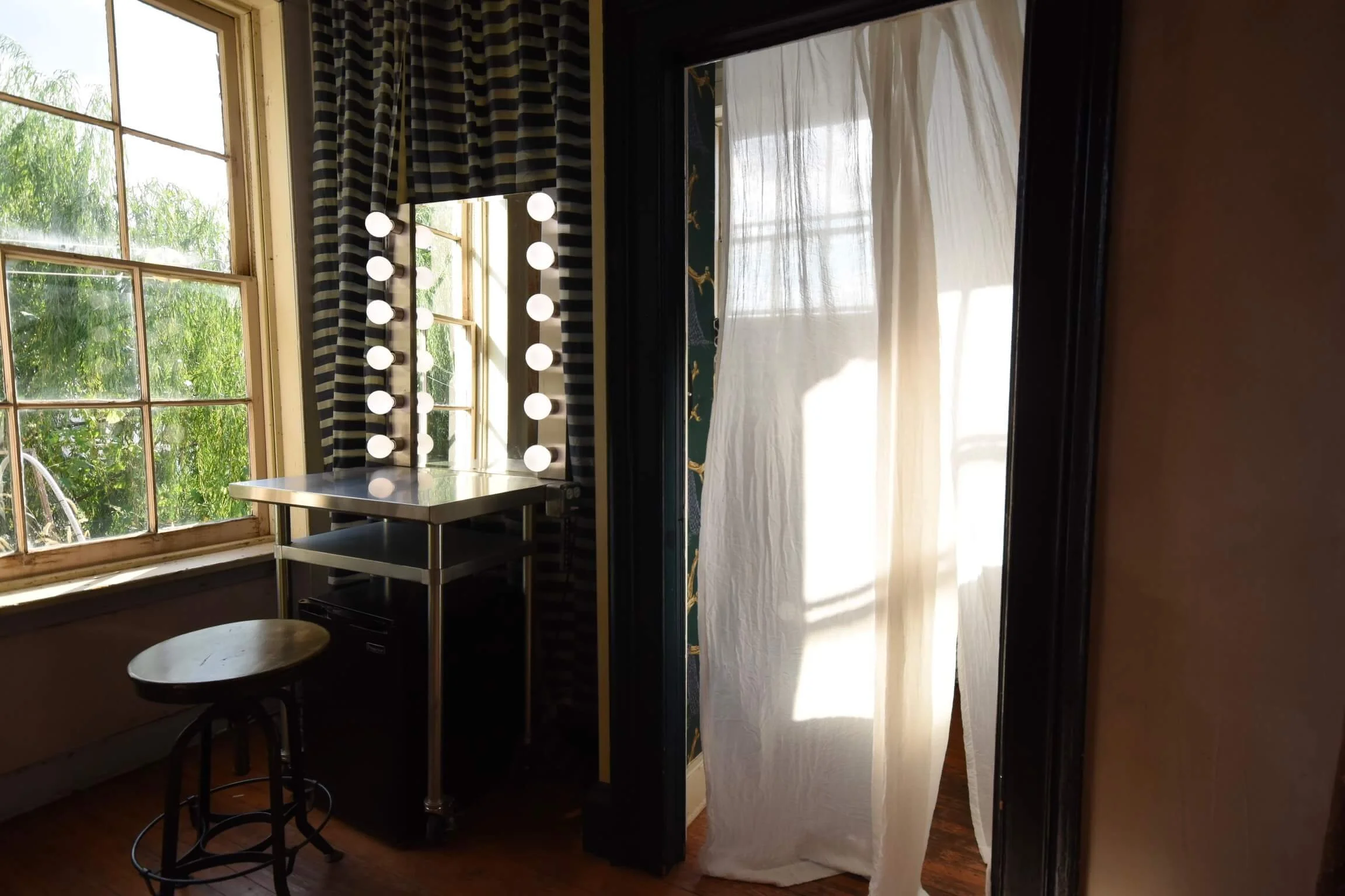 A room with a vanity mirror surrounded by lights, a window with a view of greenery, a striped curtain, a small table, and a white curtain backing a doorway.