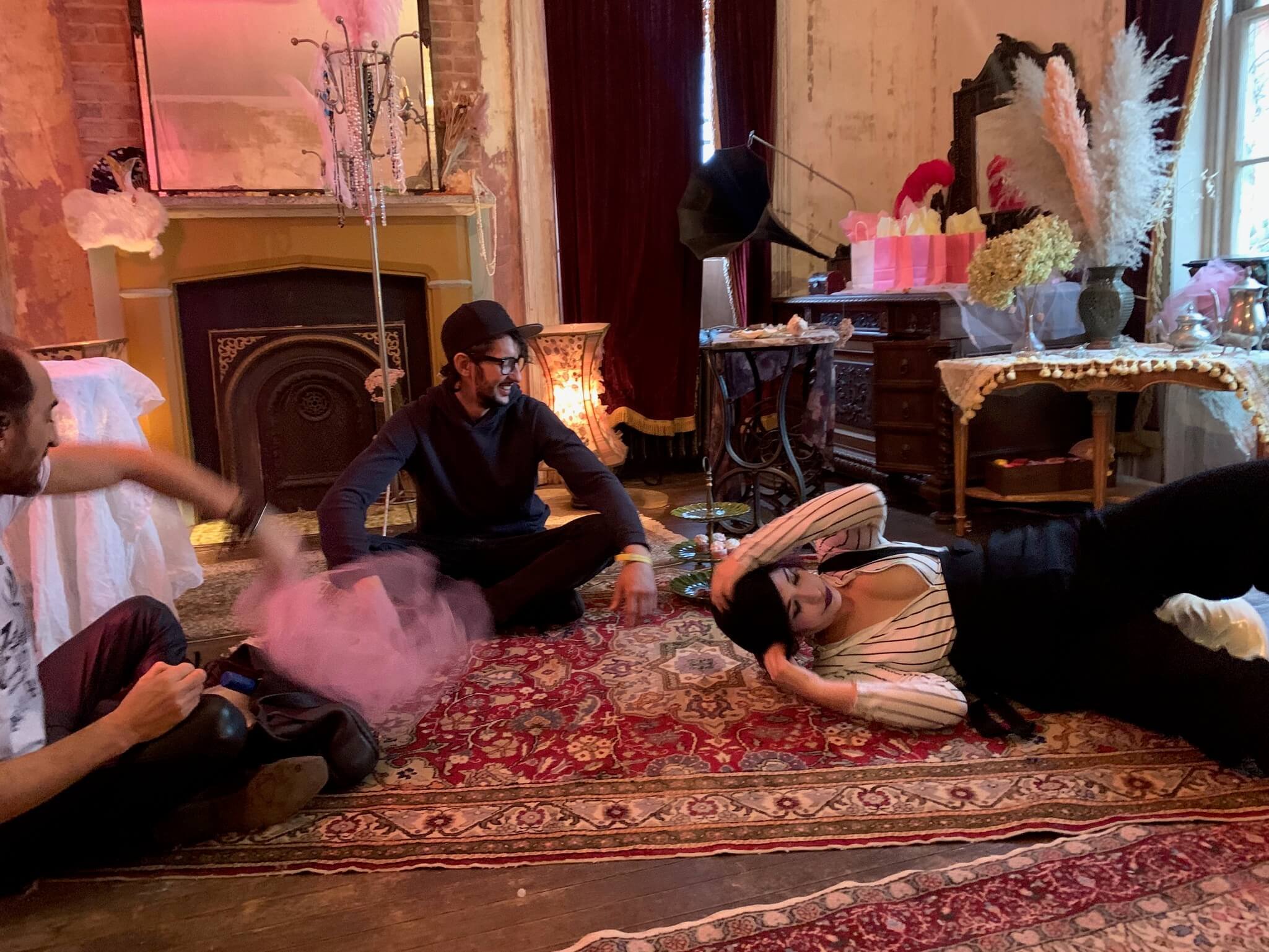 A group of people are sitting on a patterned rug in a cozy, vintage room with a fireplace and ornate furniture. One woman is lying on her side on the floor, while two men sit nearby, smiling and engaging in conversation. The room is decorated with fl