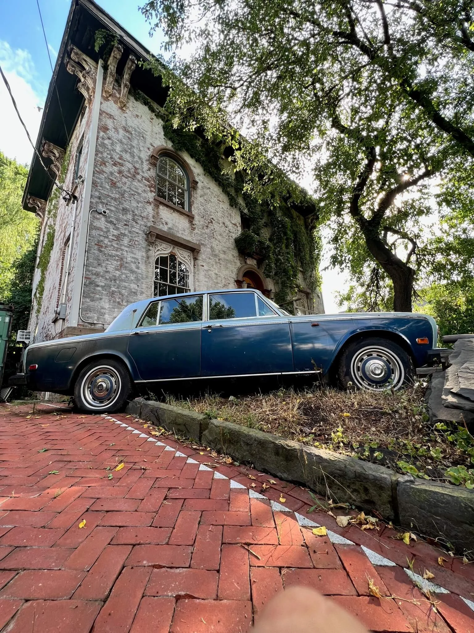 Vintage blue car parked on a brick sidewalk in front of an old brick building with ornate window frames and greenery on the walls, surrounded by trees.