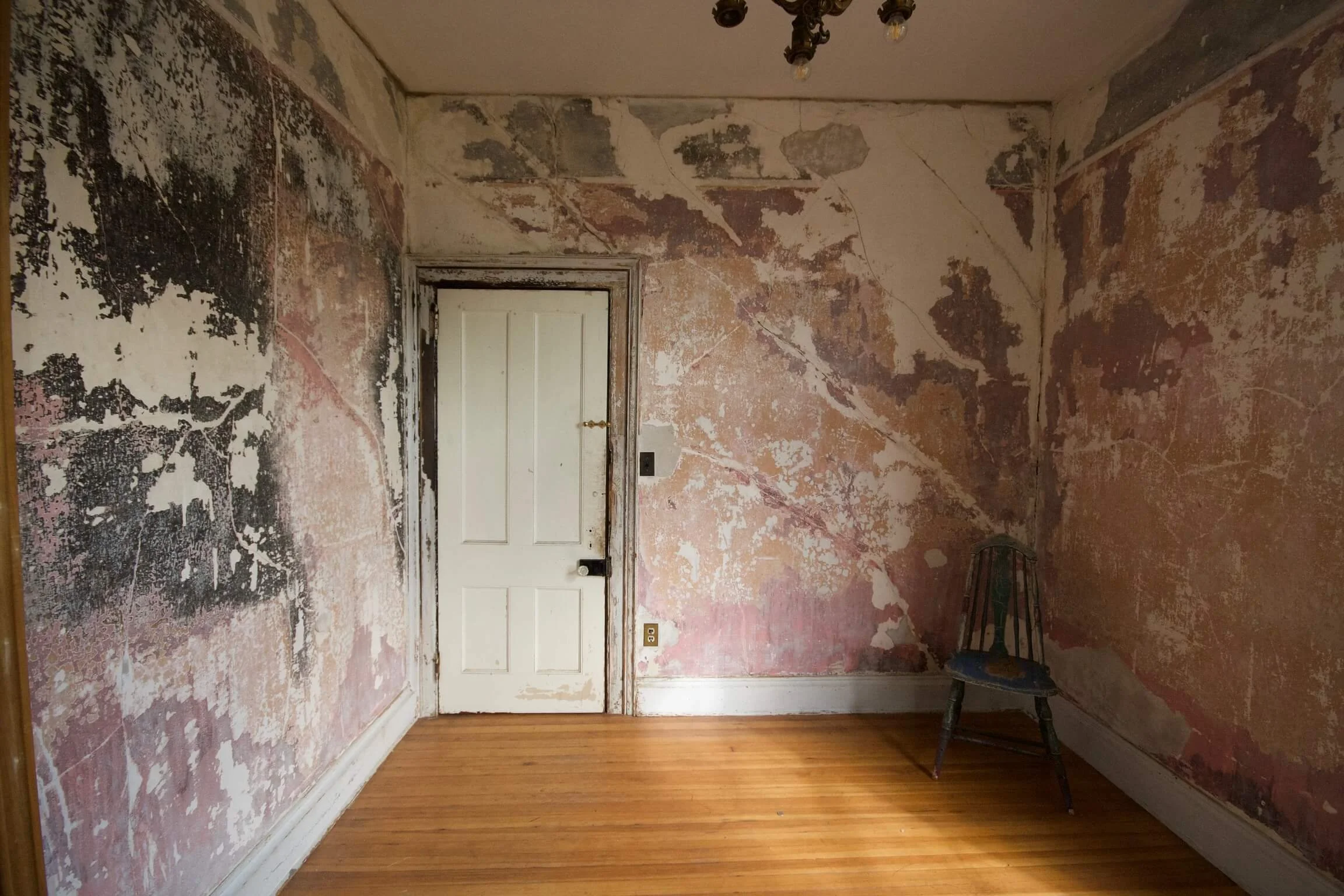 A room with peeling paint and wallpaper on the walls, a lone wooden chair in the corner, a hardwood floor, and a closed door with a doorknob. The ceiling has a small light fixture.