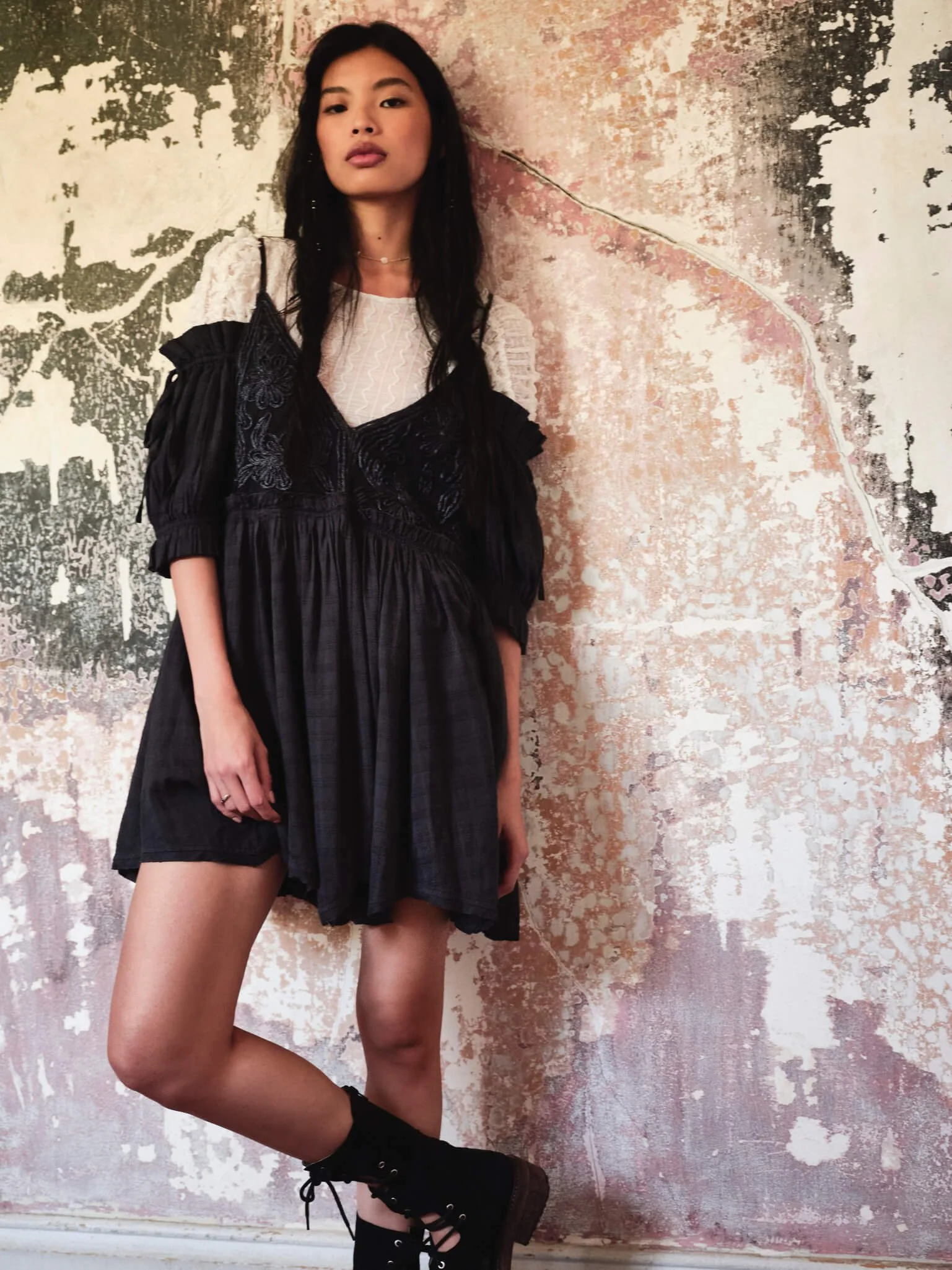 A woman with long black hair and a confident expression standing against a textured, peeling wall with pastel colors. She is wearing a layered outfit with a white lace top underneath a black dress with ruffled sleeves, paired with black combat boots.