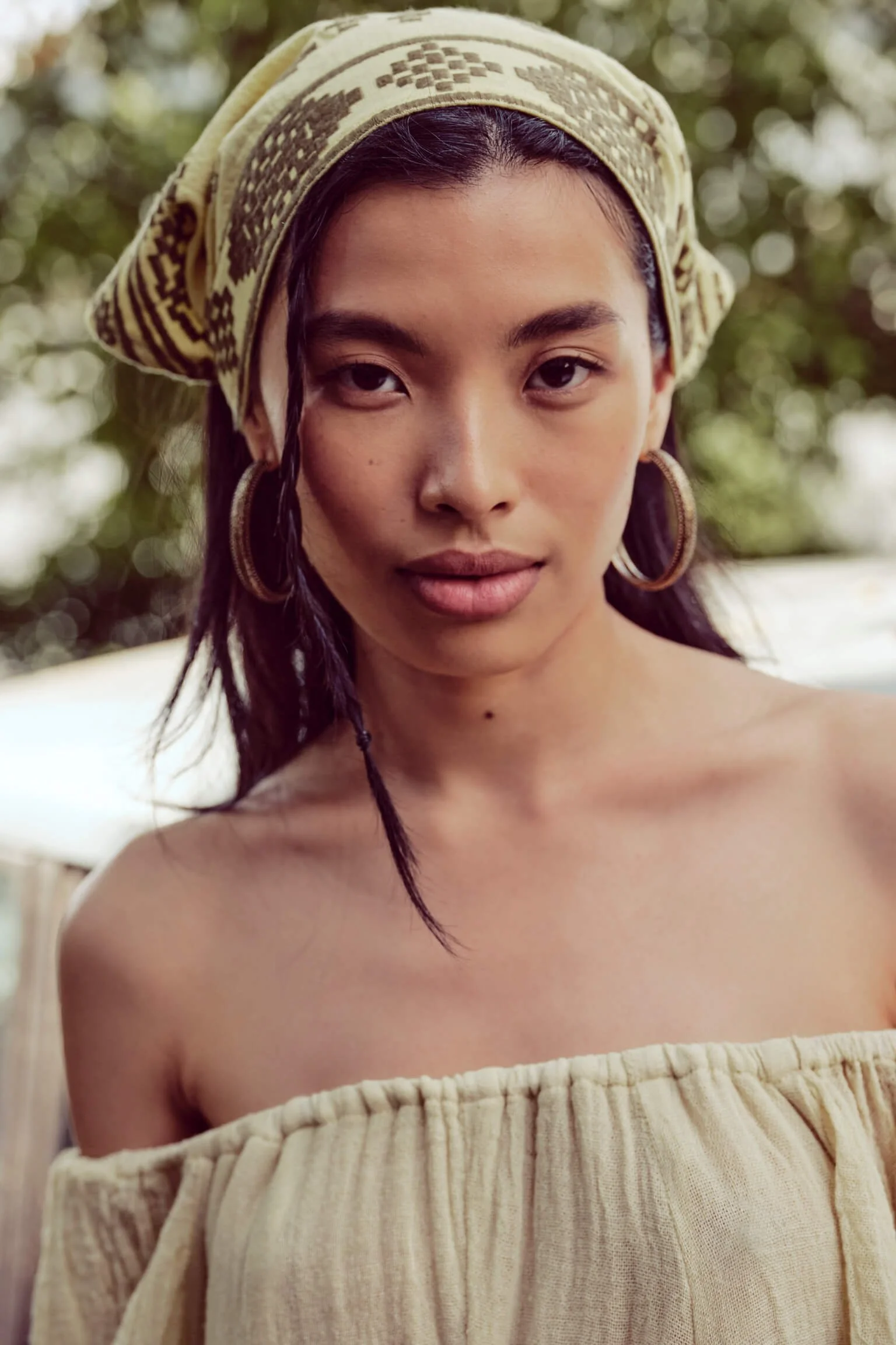 Close-up of a woman with dark hair, wearing a patterned headscarf and large hoop earrings, outdoors with blurred green foliage background.