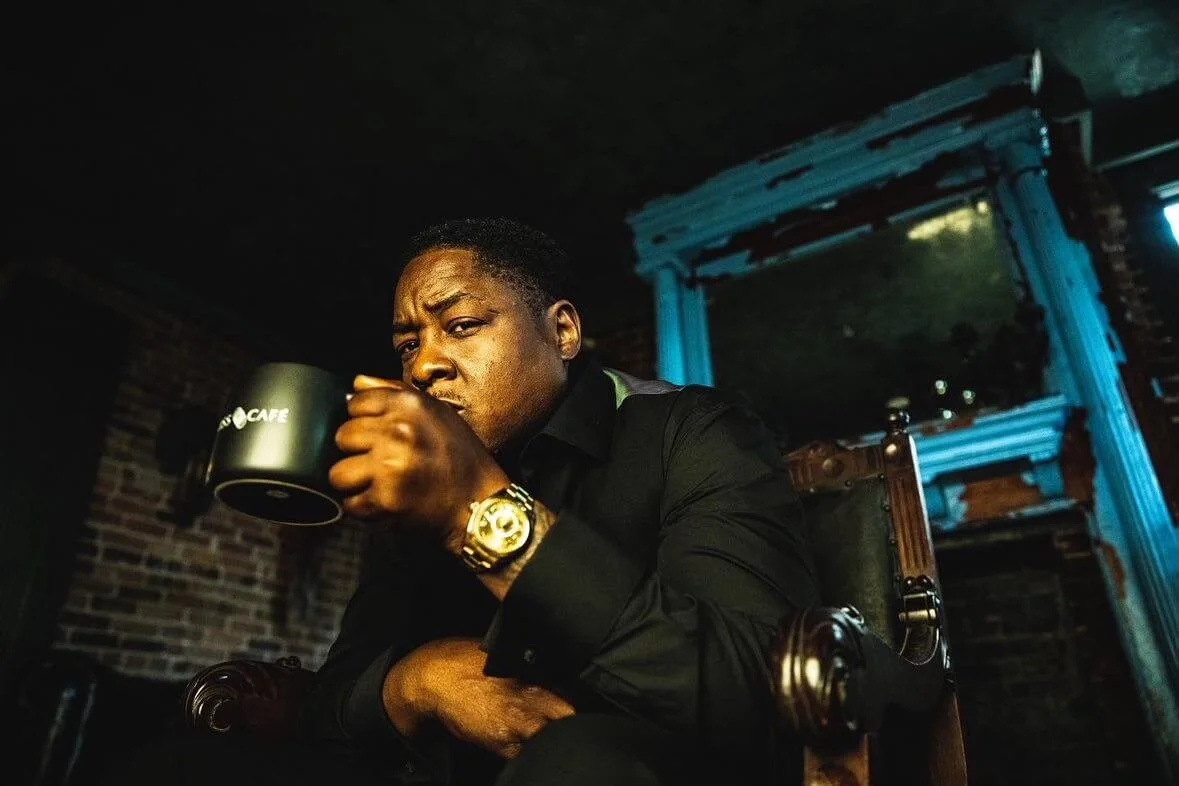 Man in black shirt sitting on wooden chair, holding a black coffee mug, in a dimly lit room with brick wall and vintage decor.
