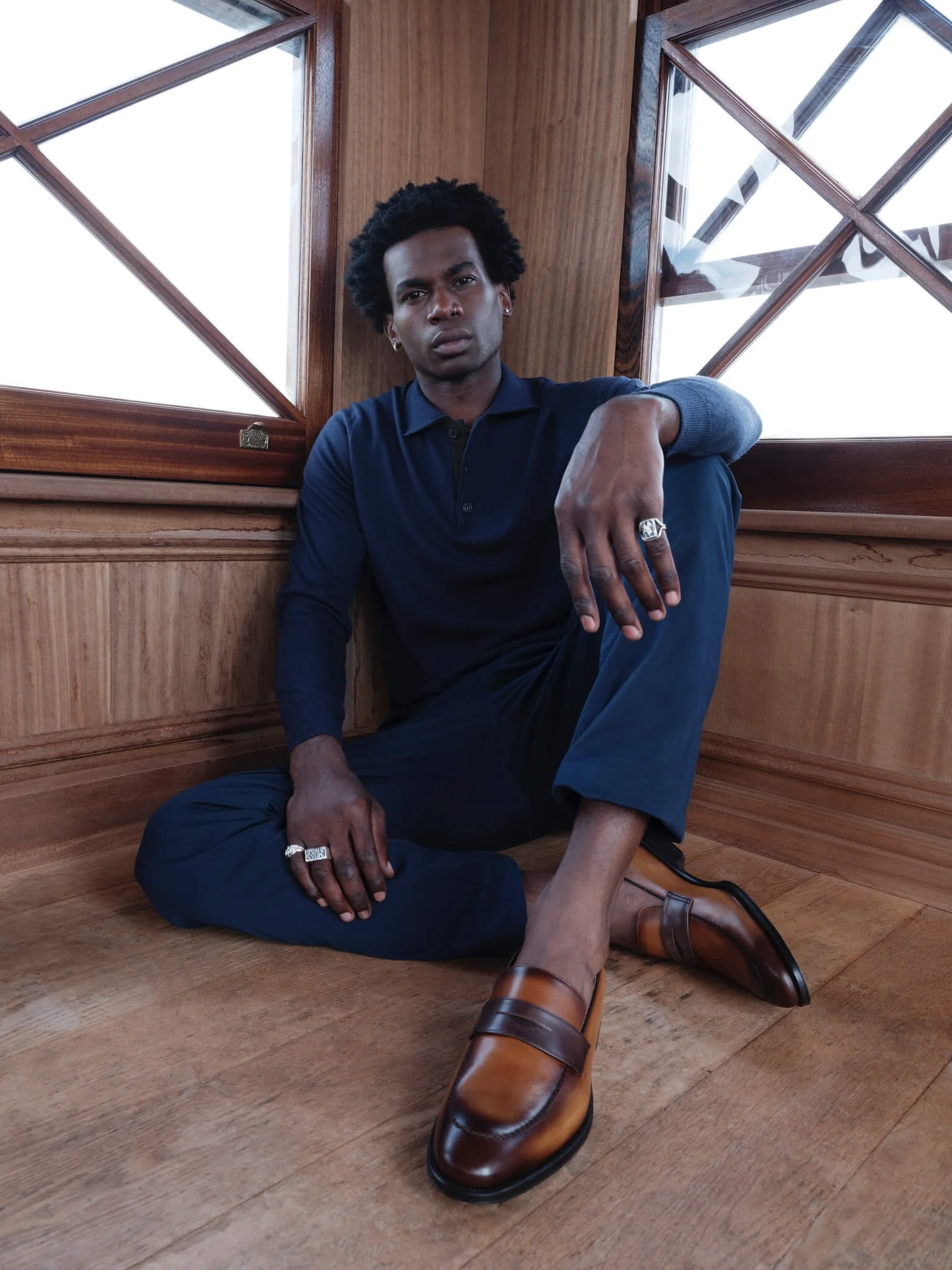 A man with curly hair sits on a wooden floor, leaning against a wooden wall near windows. He is dressed in a navy blue outfit, wearing brown leather shoes, silver rings, and earrings.