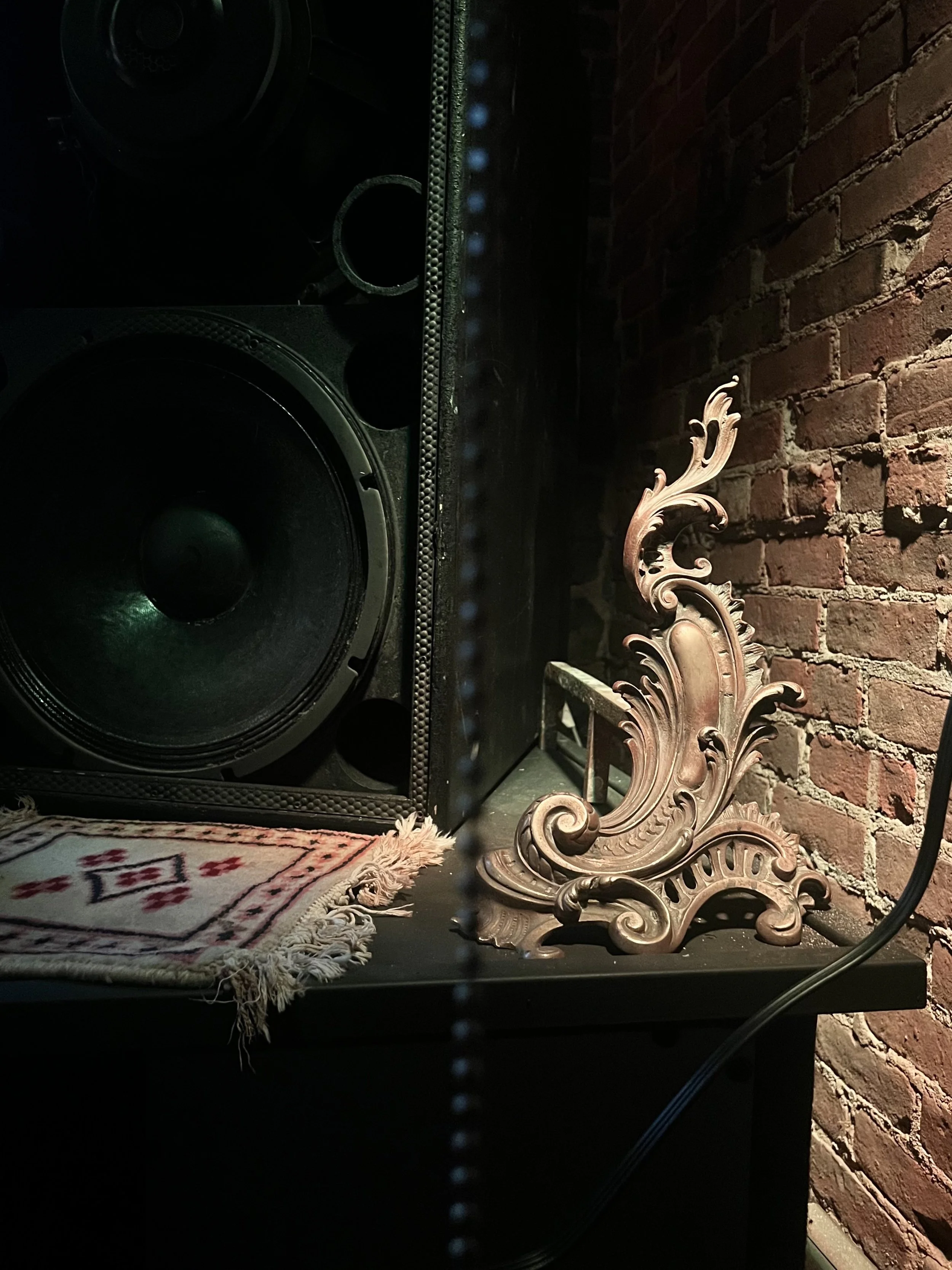 Decorative ornate bronze sculpture on a black table next to a large speaker, with a brick wall in the background and a small patterned rug partially visible.