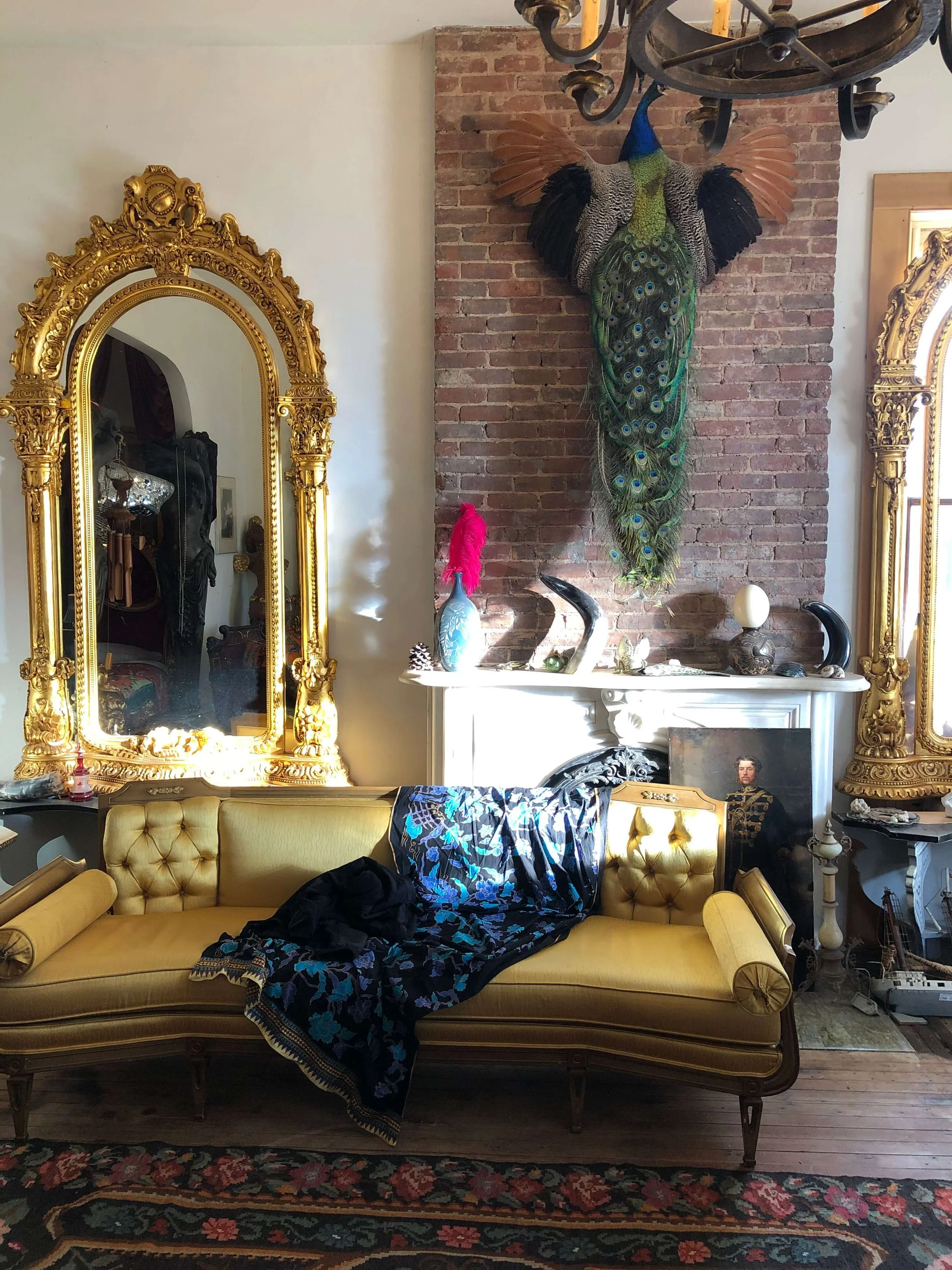 Living room with vintage yellow sofa, large ornate gold mirrors, a brick wall with a mounted peacock display, and eclectic decorations on a white fireplace mantel.