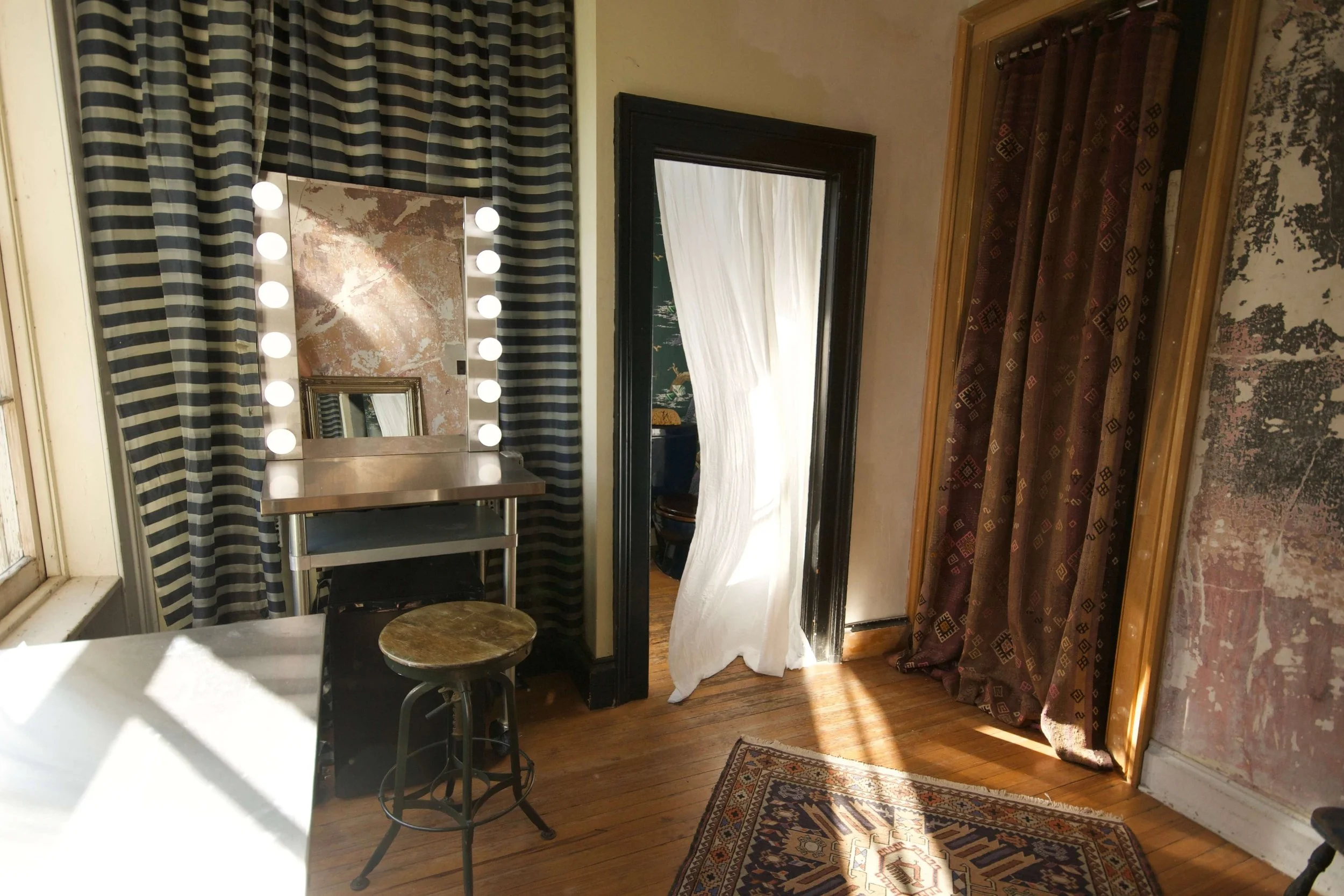 Interior of a room with a makeup mirror surrounded by light bulbs, a small stool, a table, and curtains, with sunlight coming through a window and illuminating the wooden floor.