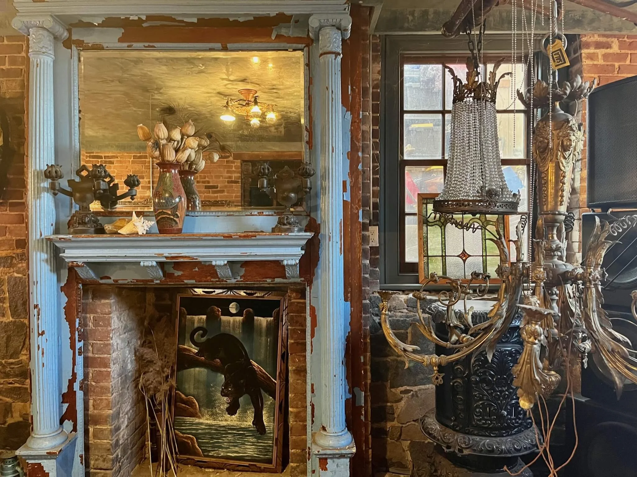 A rustic interior with a brick fireplace and an ornate mirror above it. The fireplace has a painting of a black cat leaping over water and is decorated with natural items like dried flowers and shell ornaments. To the right, there is a window with a 