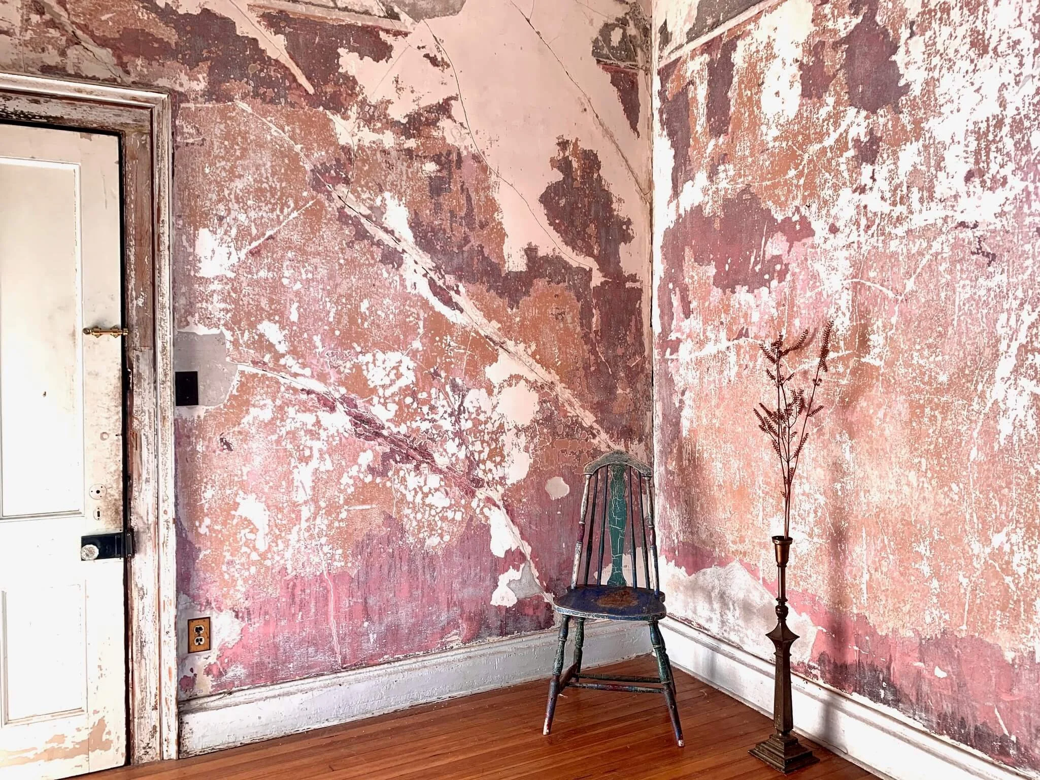 A room with peeling pink and white paint on the walls, a wooden floor, an old painted wooden chair, and a tall planter with a dried plant.