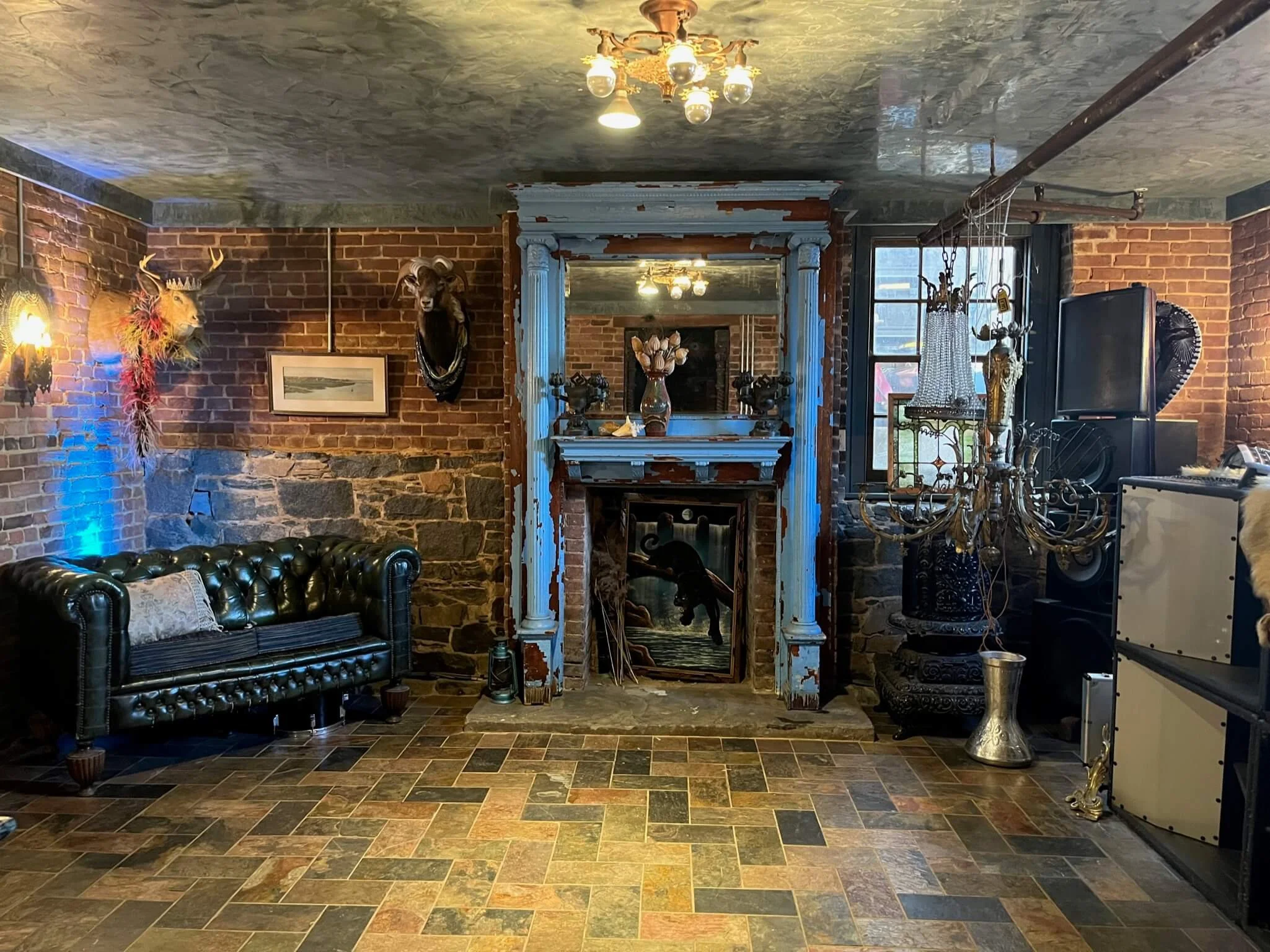 A rustic living room with exposed brick walls, a stone fireplace, and a stained glass window. Decorations include a mounted deer head, a mounted ram head, a framed landscape on the wall, a chandelier hanging from the ceiling, and a black leather tuft