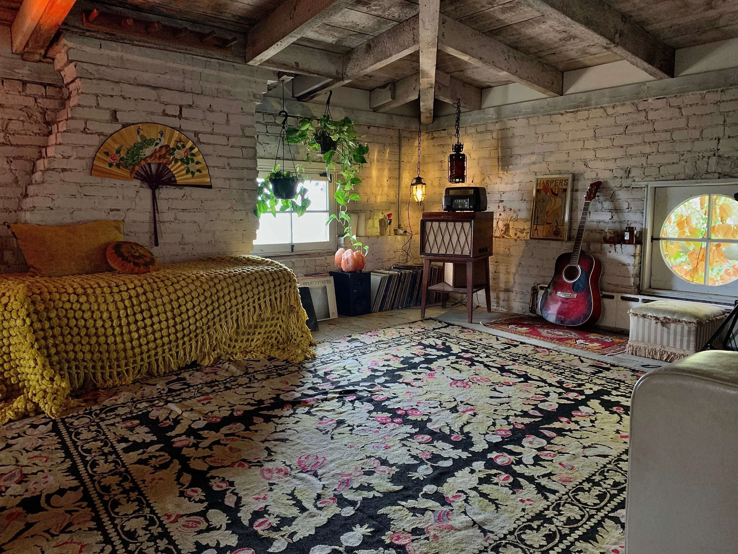 Living room with white brick walls, a patterned floral rug, a yellow crocheted blanket on a couch, vintage decor, a guitar, and potted plants near a window.