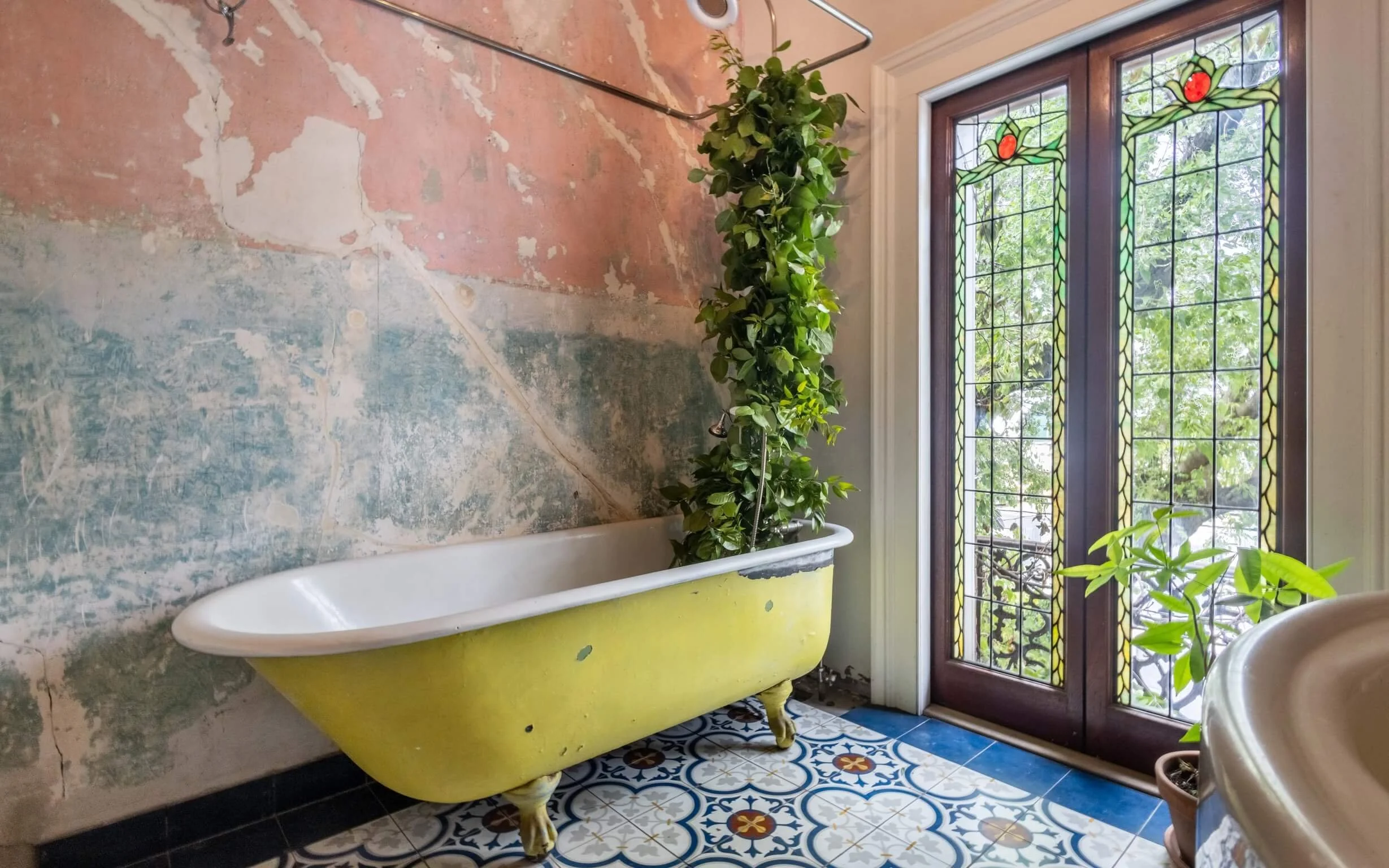 Vintage yellow clawfoot bathtub with green potted plants, next to stained glass window with greenery outside, and a weathered wall with peeling paint.