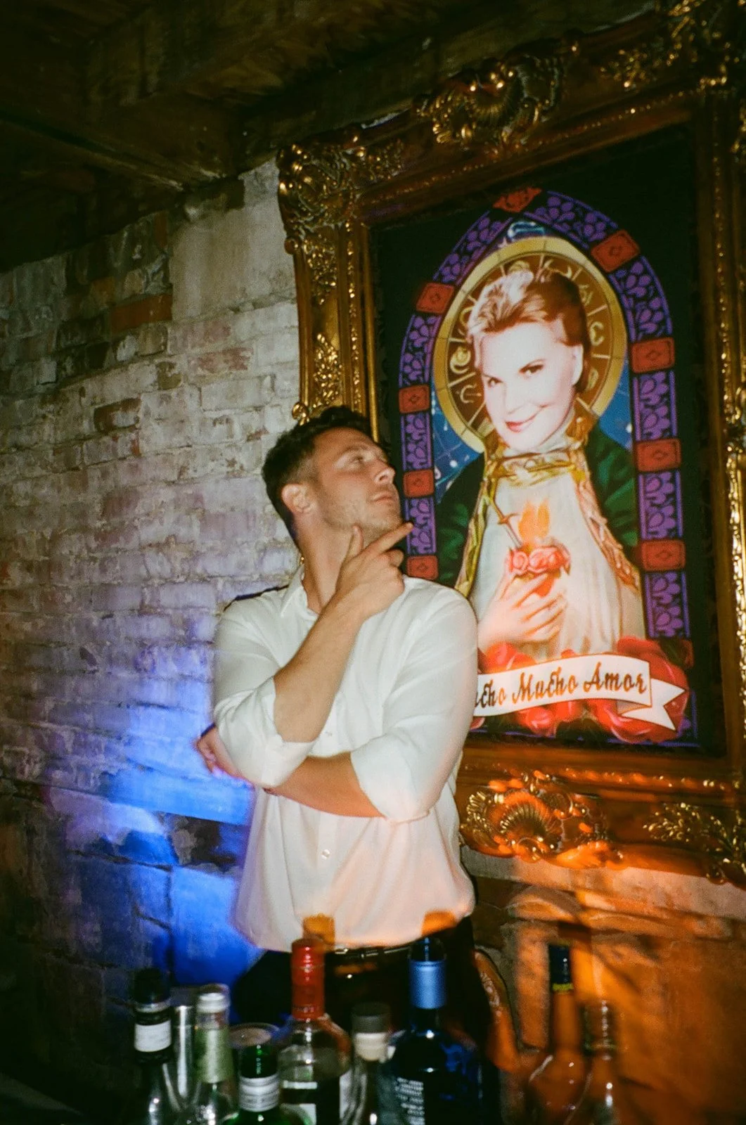 A man in a white shirt stands in front of a colorful religious portrait framed in gold, with a brick wall behind him. The man is contemplating the picture, with one hand on his chin and looking up. Several bottles of alcohol are on the table in front