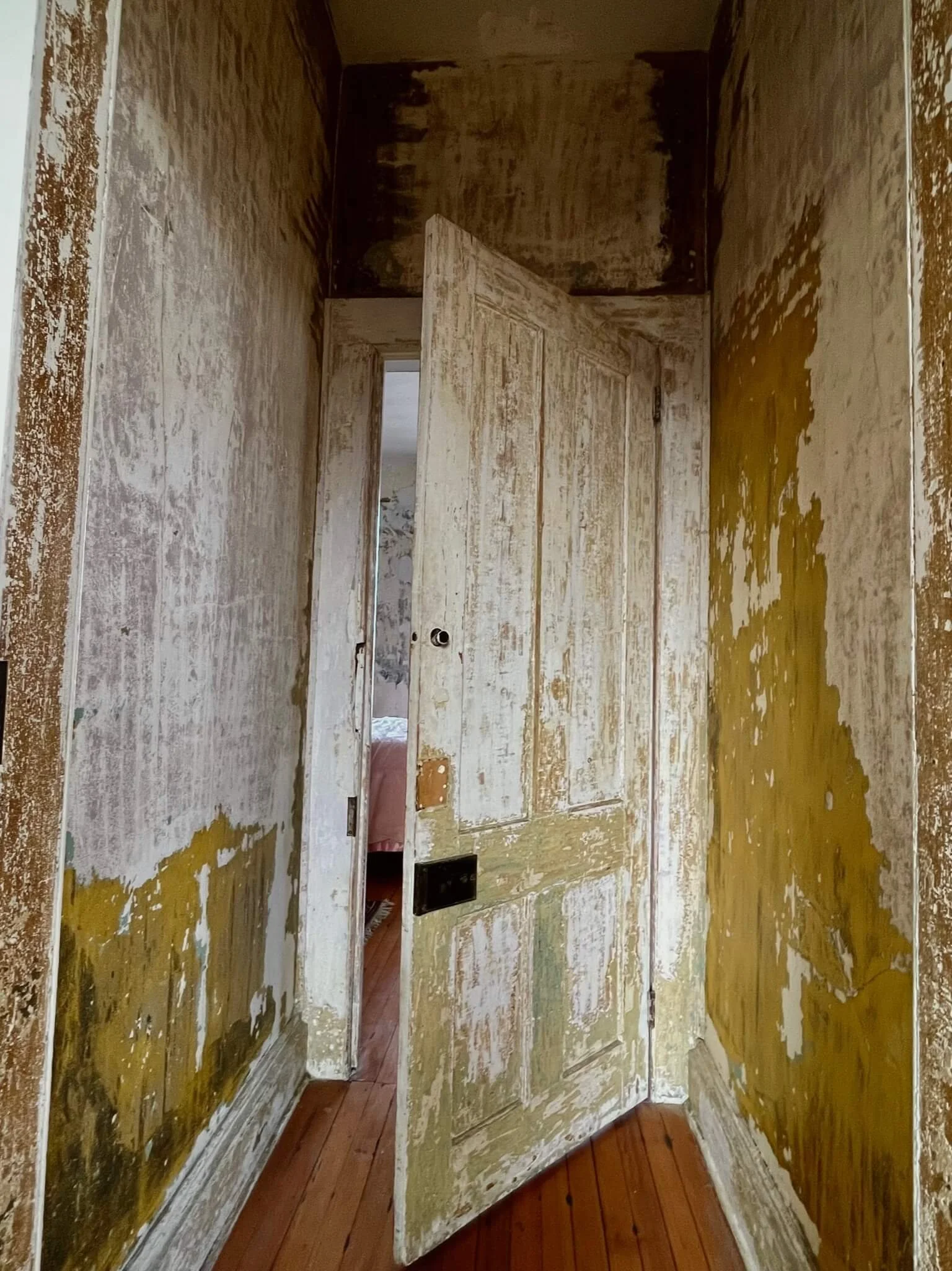 An old, weathered door slightly ajar in a hallway with peeling paint on the walls, revealing a room with a bed in the background.