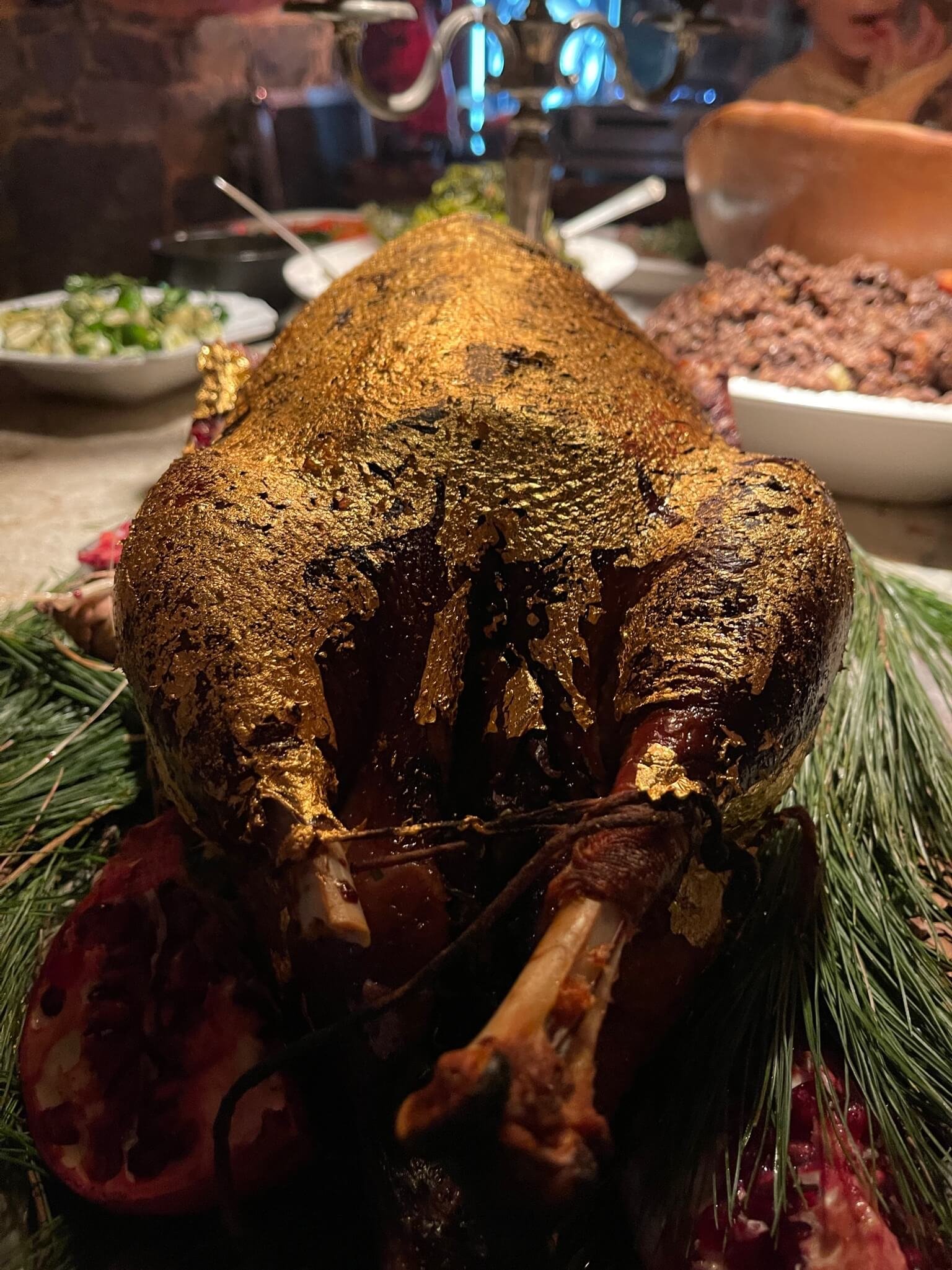 Roast turkey with golden brown crispy skin, garnished with pine branches, surrounded by pomegranate and additional food dishes in the background.