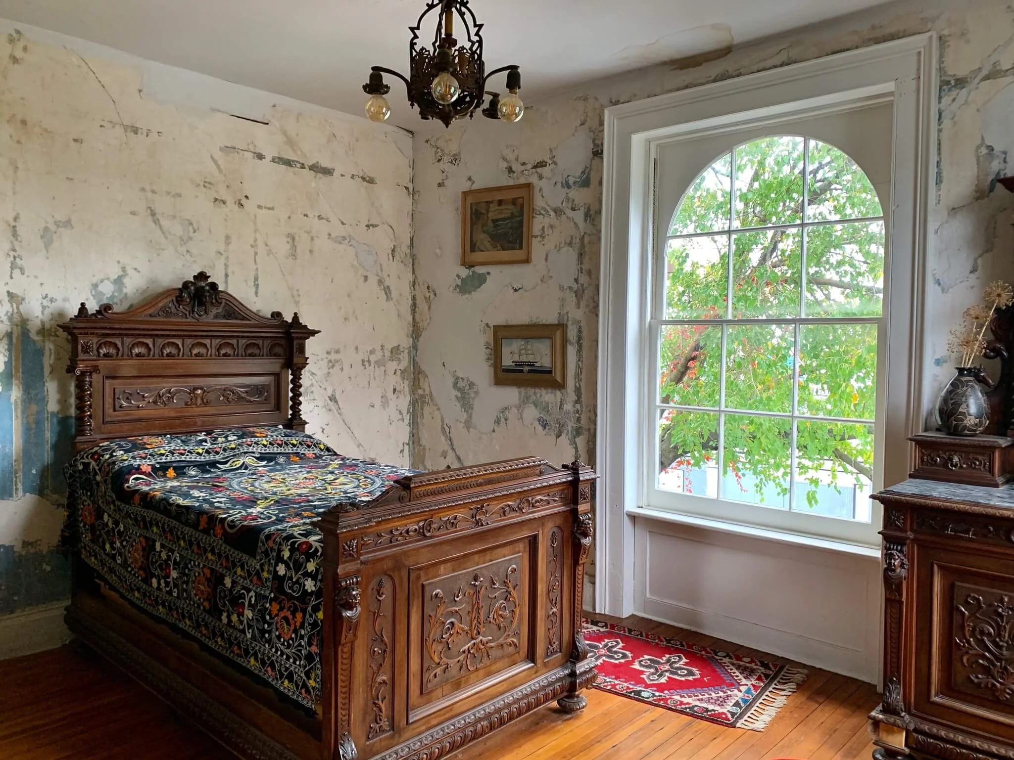 A vintage bedroom with antique wooden furniture, a carved bed, a dresser with decorative items, a colorful embroidered bedspread, a window showing green trees outside, and a chandelier hanging from the ceiling.