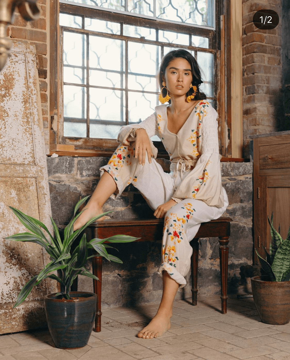 A woman wearing a cream-colored embroidered jumpsuit sits on a wooden bench by a window in a rustic interior with brick and stone walls, with potted plants nearby.