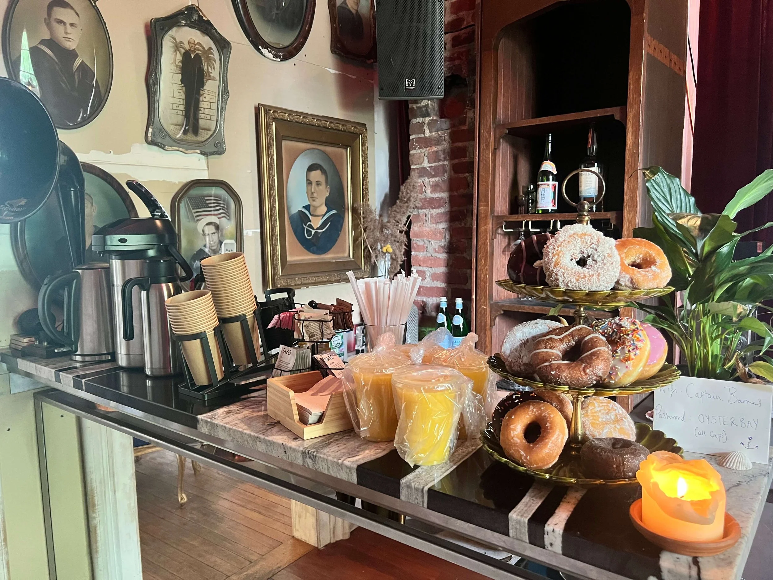 Coffee and tea station with stacked cups, honey, and a three-tiered tray of assorted donuts in a cozy cafe with vintage framed photos on the wall and a candle.