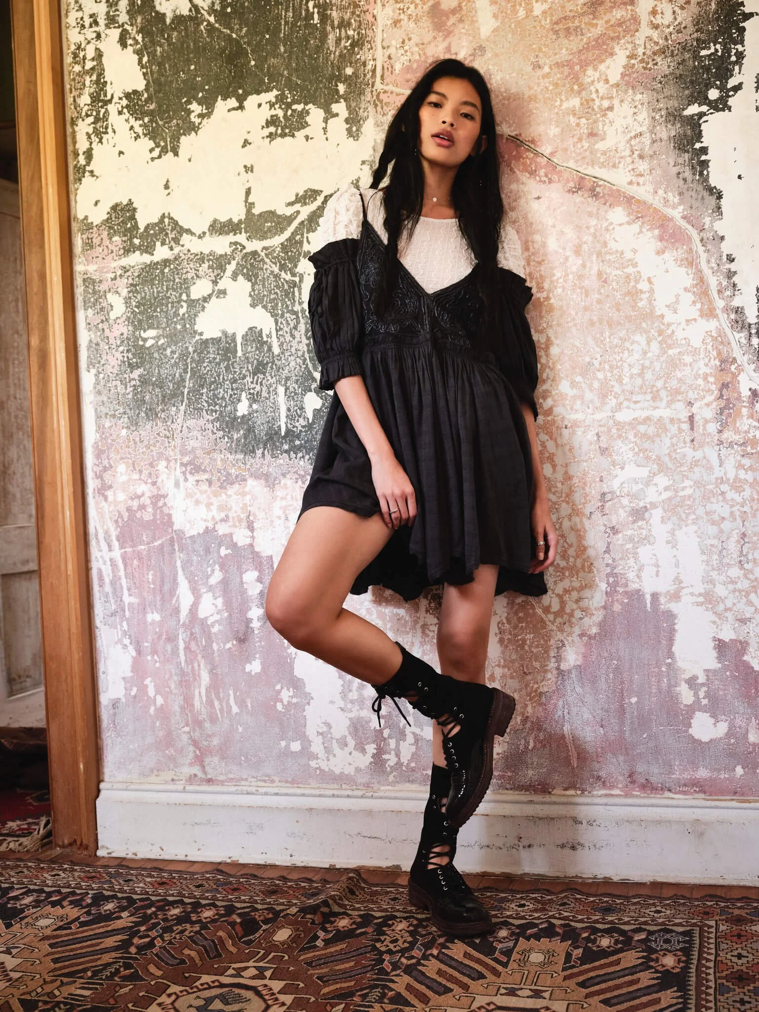 Young woman in black dress and combat boots standing against a textured wall.