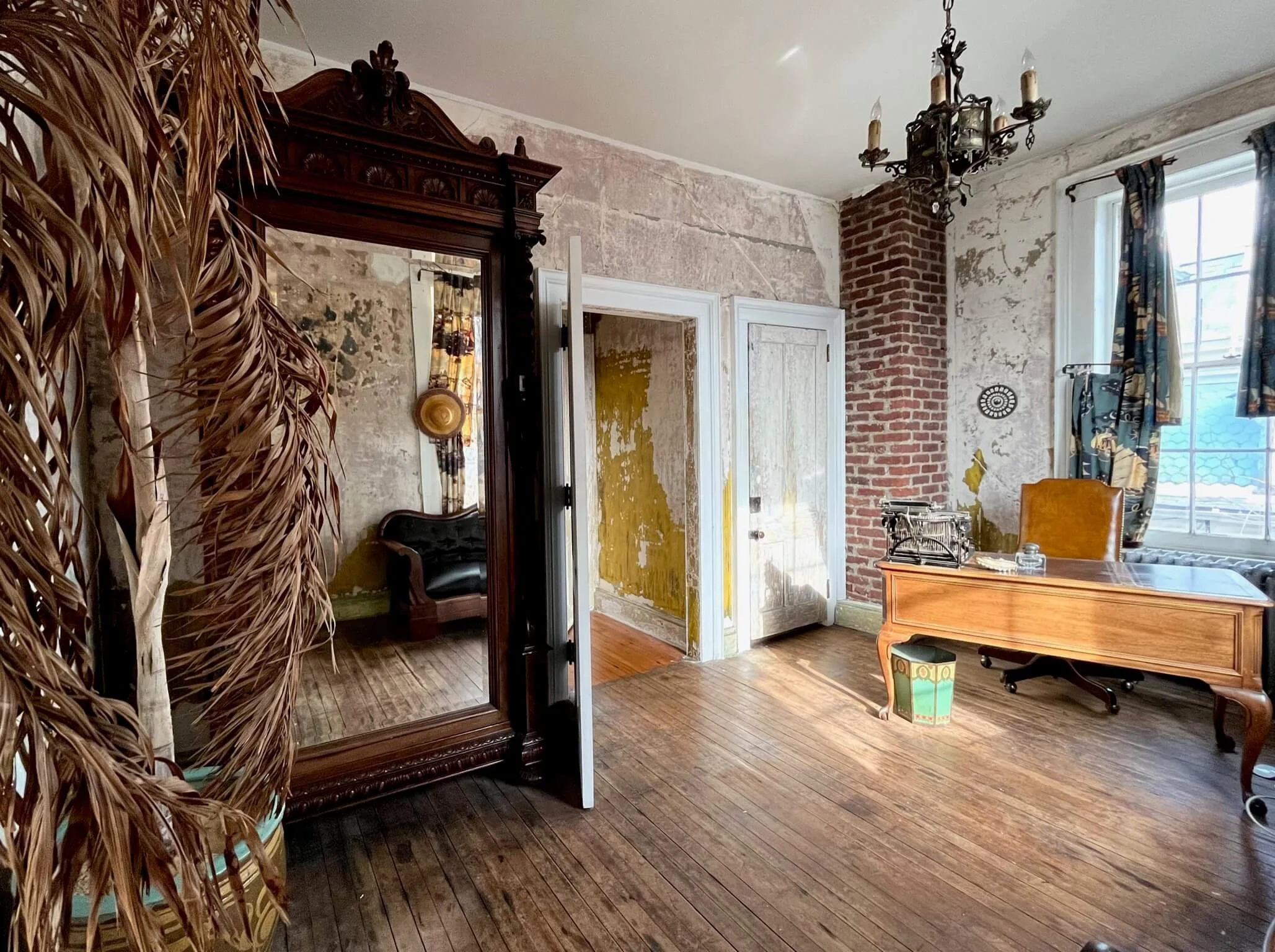 Interior of a vintage-style room with wooden floors, exposed brick and distressed wall surfaces, a large ornate mirror, a wooden desk with a leather chair, a window with patterned curtains, and a chandelier hanging from the ceiling.