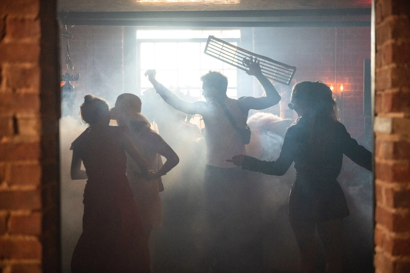 Silhouettes of people dancing and socializing in a dimly lit room with smoky atmosphere, backlit by a window.