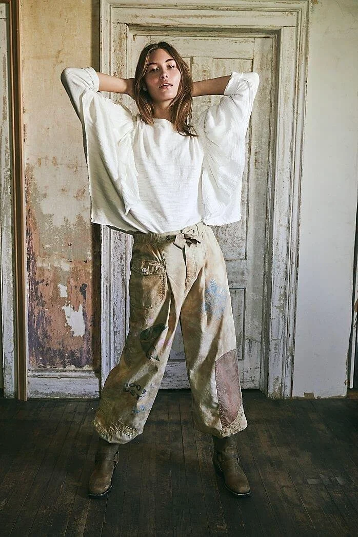 A woman with brown hair wearing a loose white top and paint-stained beige work pants, standing with hands behind her head in front of a worn, distressed door.