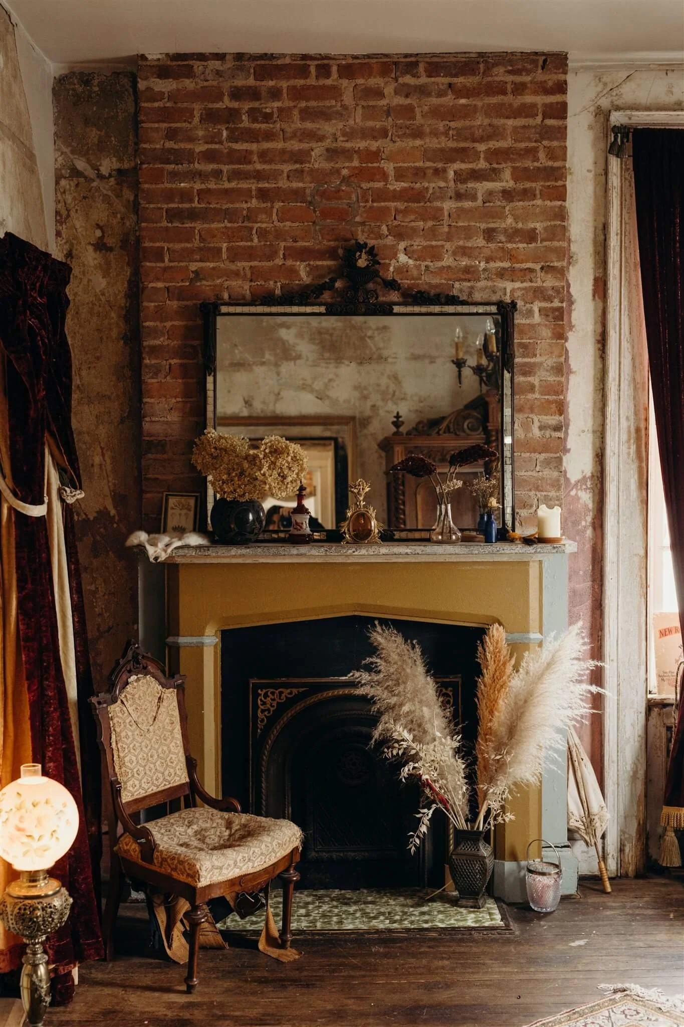 Vintage inspired living room with exposed brick wall, ornate mirror, dried flowers on fireplace mantel, vintage chair, pampas grass in vase, candle, and warm ambient lighting.