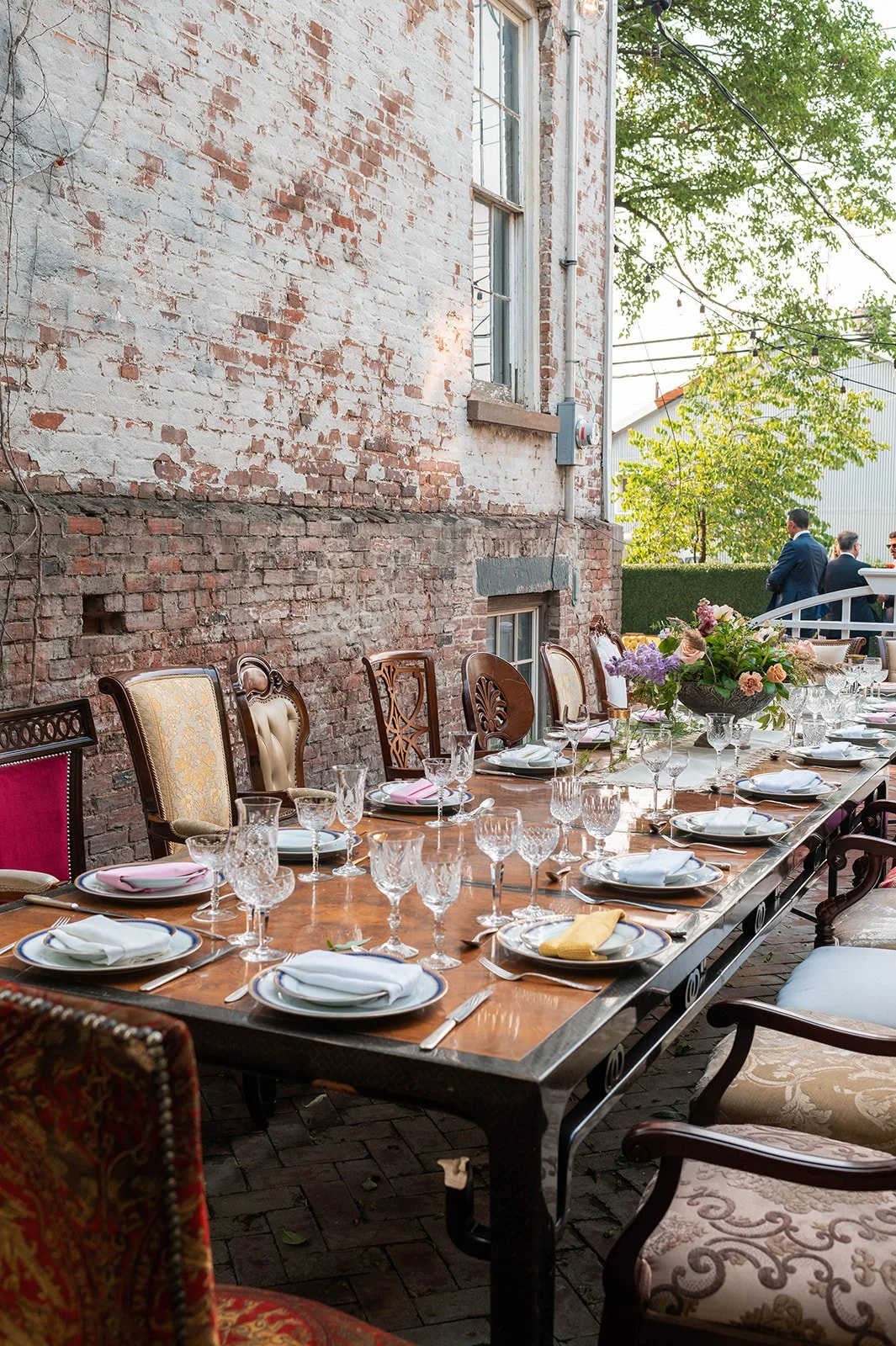 A long dining table set for a meal with plates, silverware, and glassware, decorated with a flower arrangement in an outdoor patio with brick wall and trees.