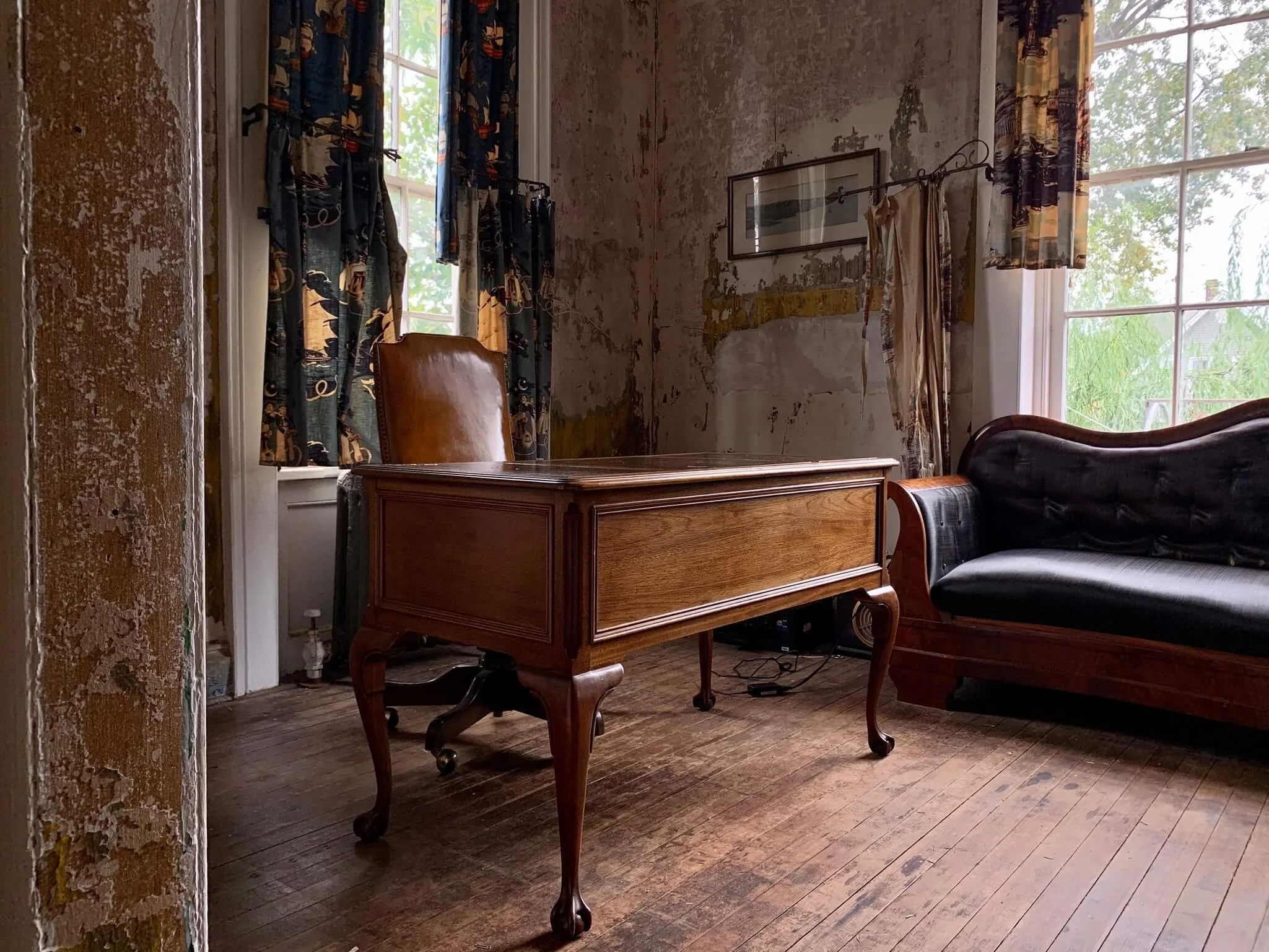 Interior of a room with wooden flooring, two large windows with vintage curtains, an antique wooden desk with curved legs, a brown leather office chair behind the desk, and a black leather upholstered sofa. The walls are partially exposed with peelin