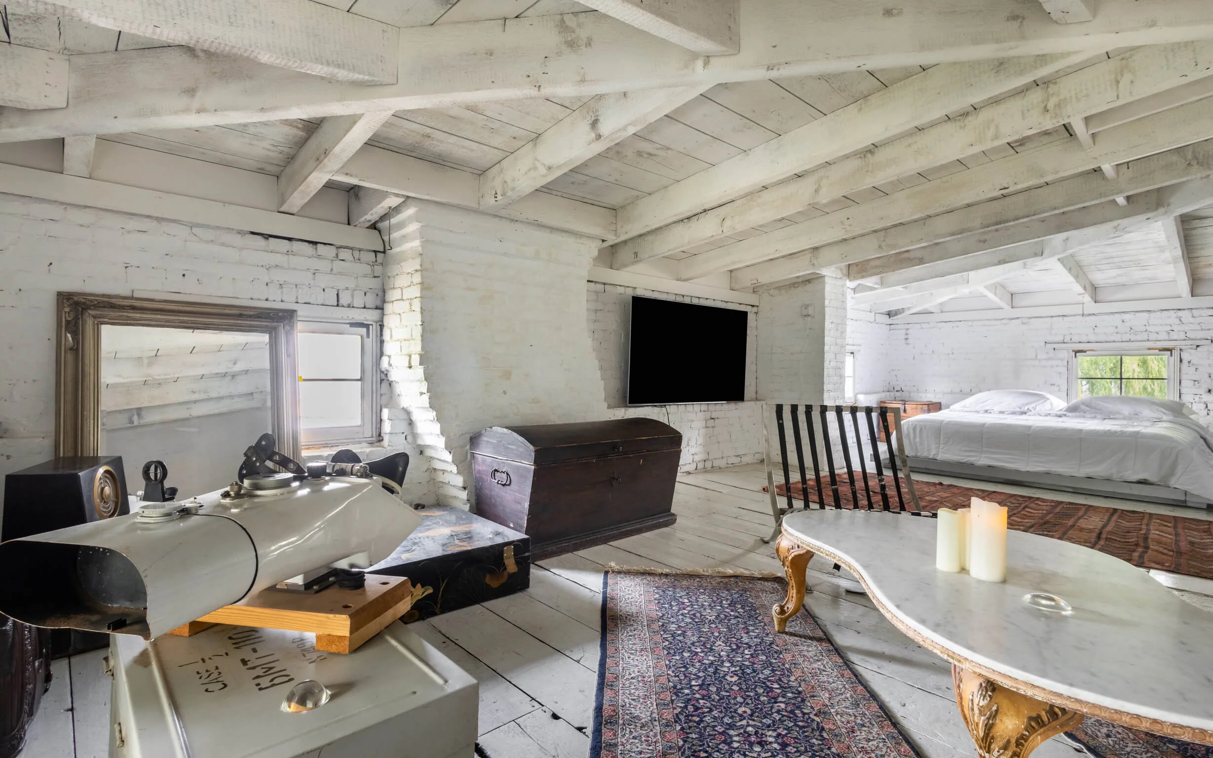 A spacious bedroom with white brick walls and ceiling beams, a large bed, a flat-screen TV, a vintage wooden chest, a decorative table with candles, and a window with natural light.