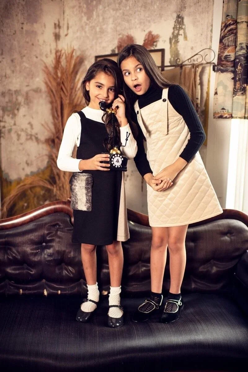 Two young girls standing on a vintage sofa, one holding a black rotary phone to her ear, in a room with rustic decor and a window with curtains.