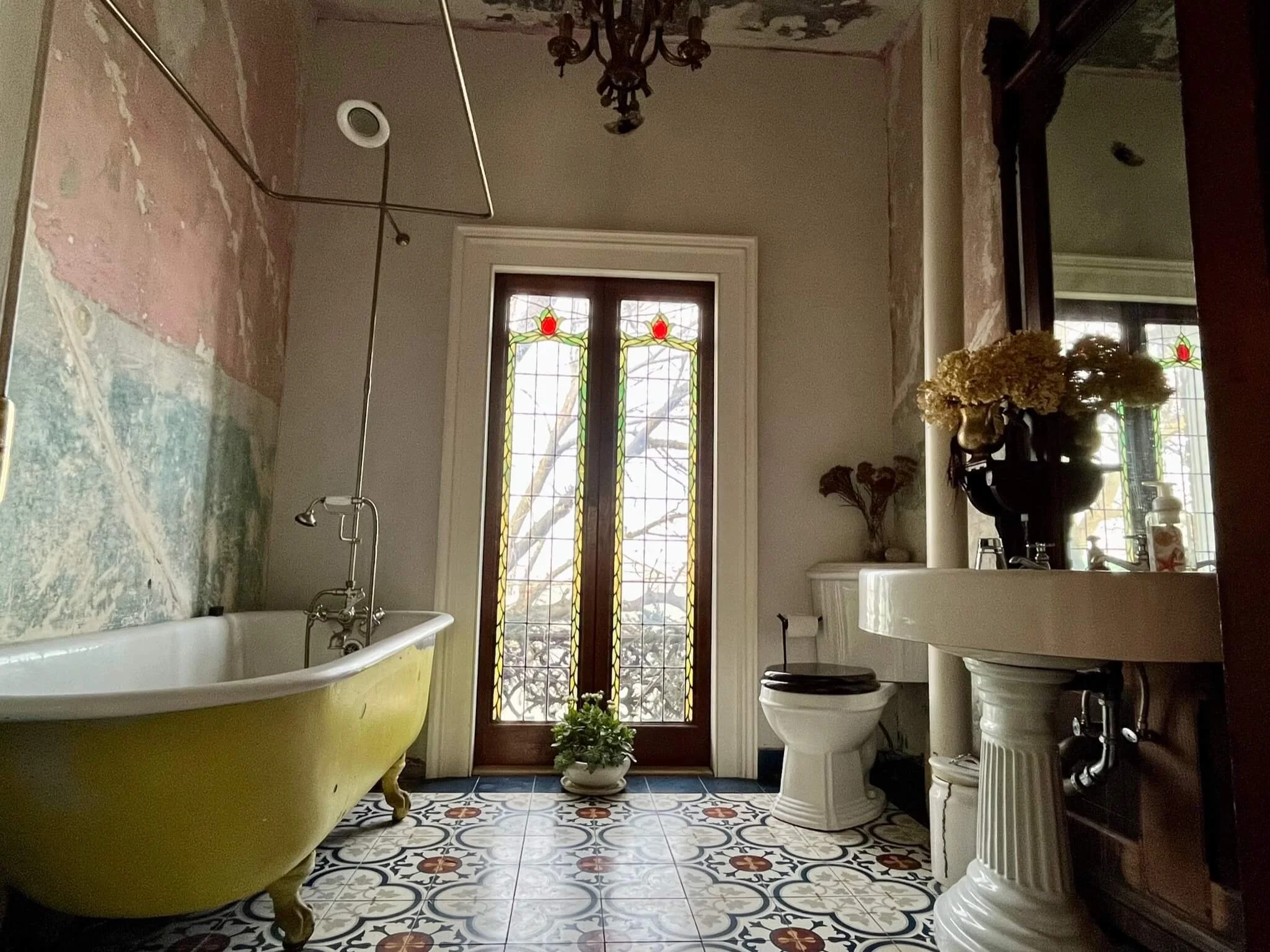 Vintage yellow clawfoot bathtub with old wall plaster, stained glass door, floral tile floor, and antique-style white pedestal sink with a dark mirror and flower arrangement.
