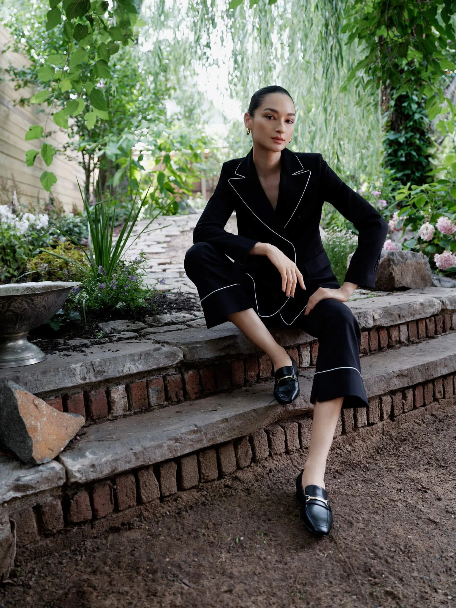 A woman dressed in a black suit with white piping sits on stone steps in a garden with lush green plants and flowers.