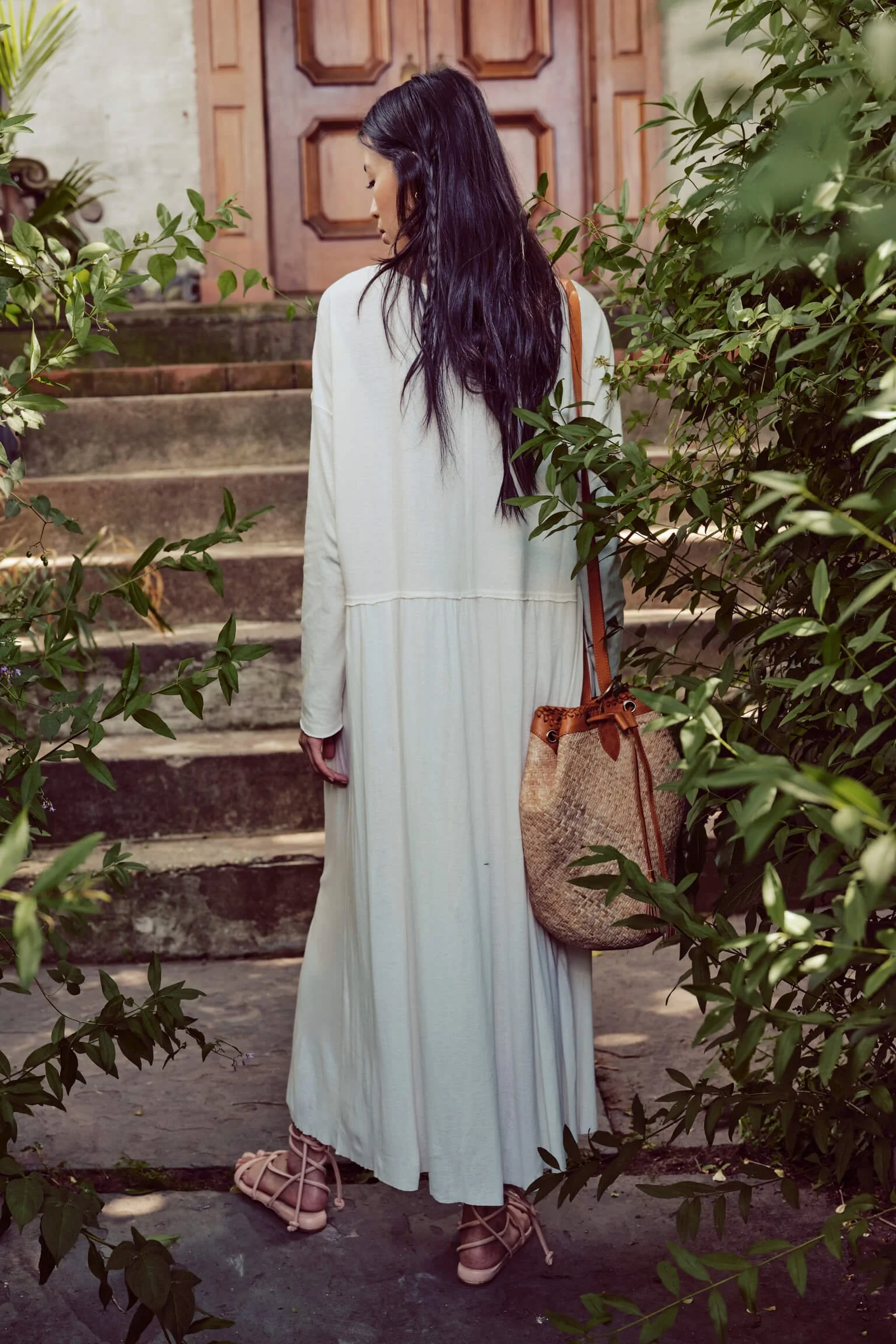 Woman in a white dress standing on stairs, surrounded by green foliage, holding a woven handbag.