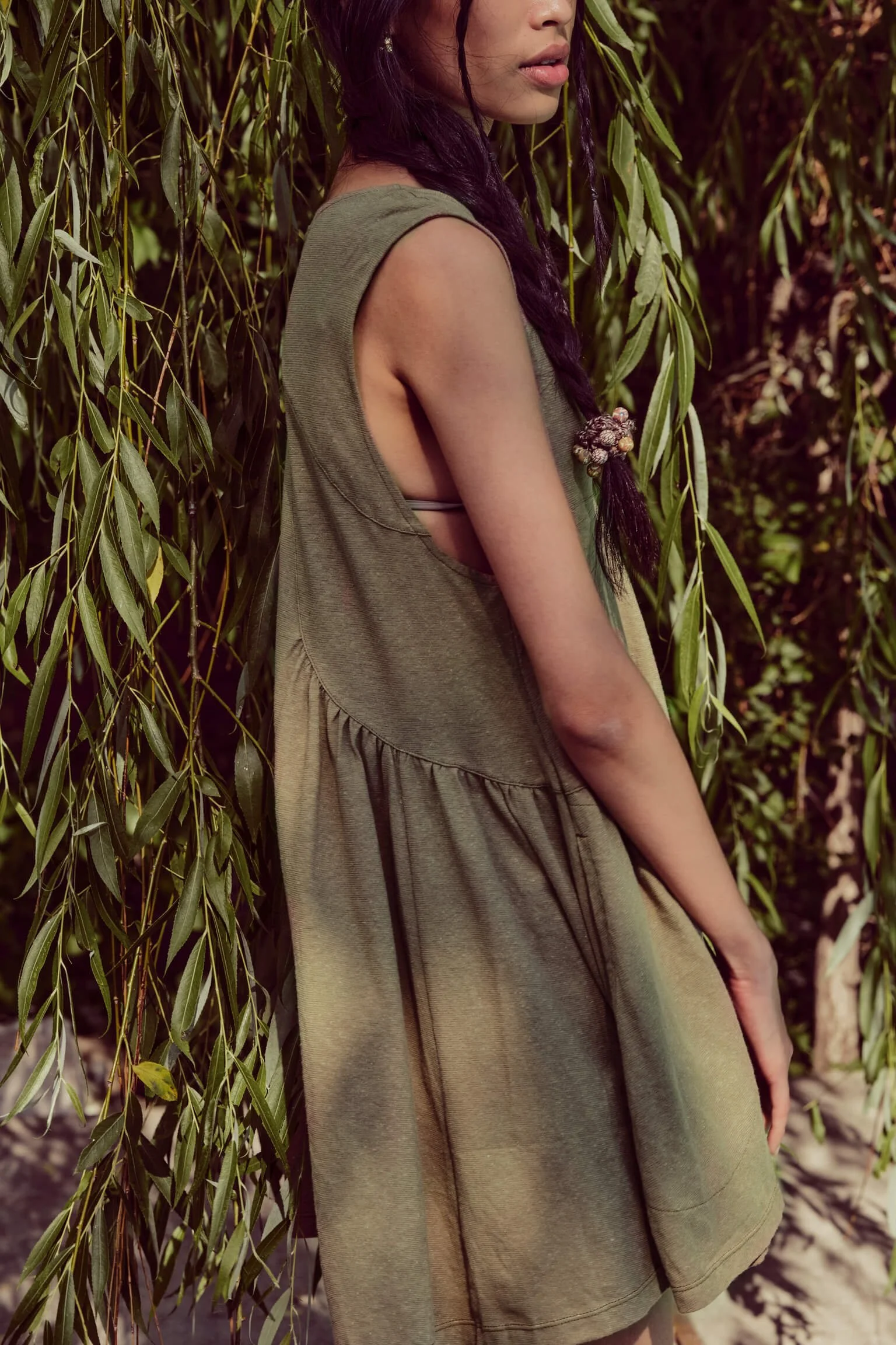 A woman wearing a loose green dress with an open armhole, standing among hanging green leaves with long, dark hair partially braided and decorated with beads.