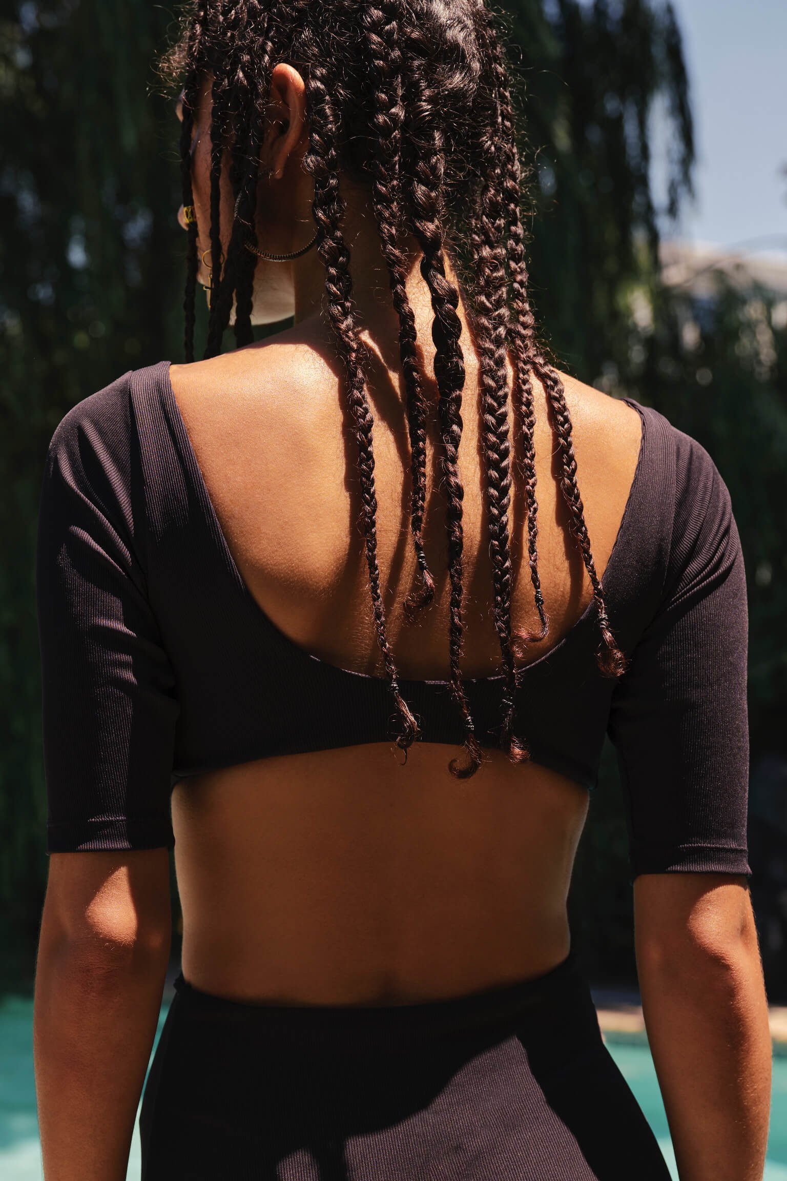 Back of a woman with long braided hair, wearing a black top with three-quarter sleeves and a black skirt outdoors on a sunny day.