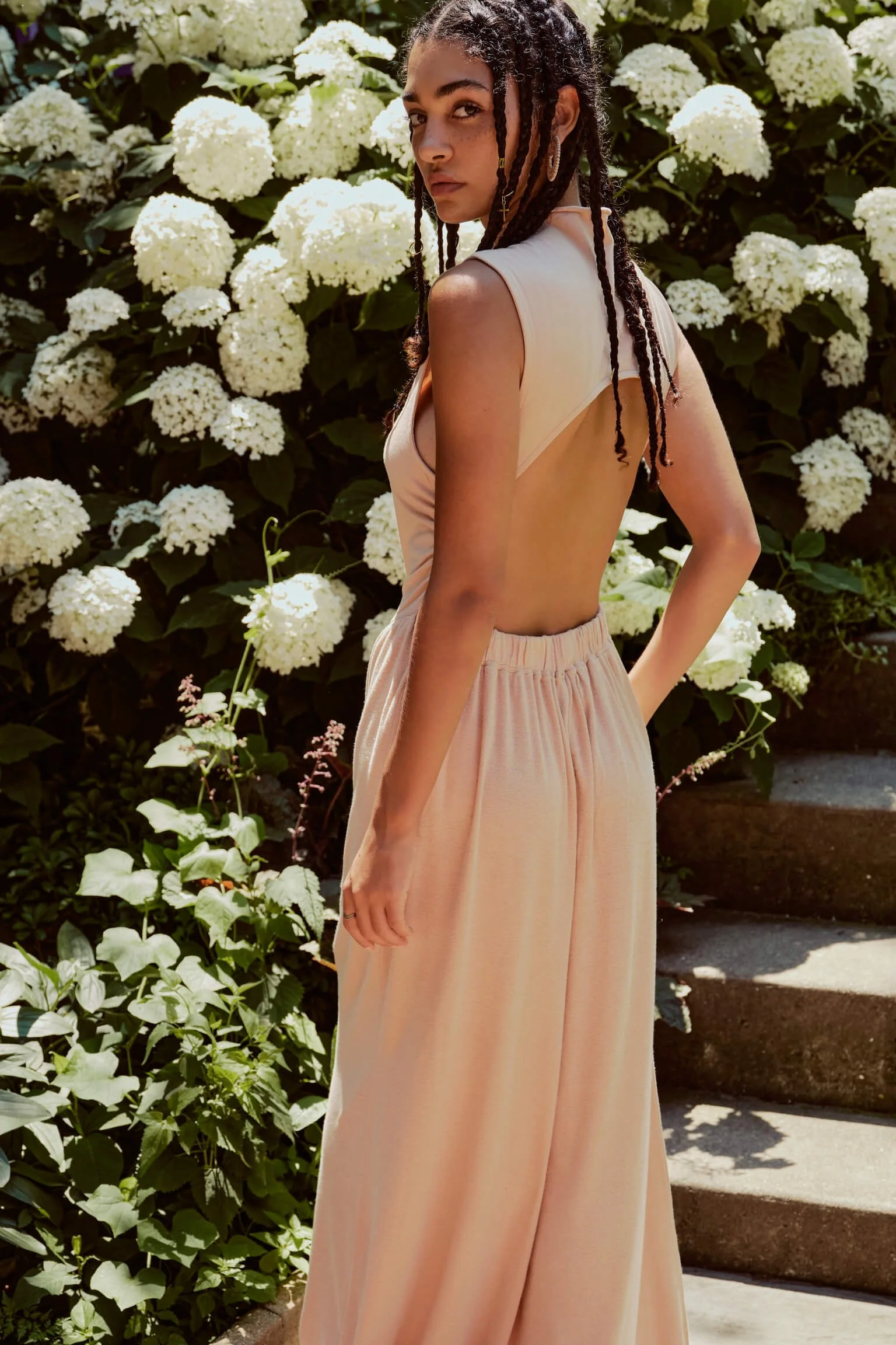 A woman with dreadlocks wearing a backless beige top and wide-leg pants standing next to white hydrangea flowers and green foliage outside.