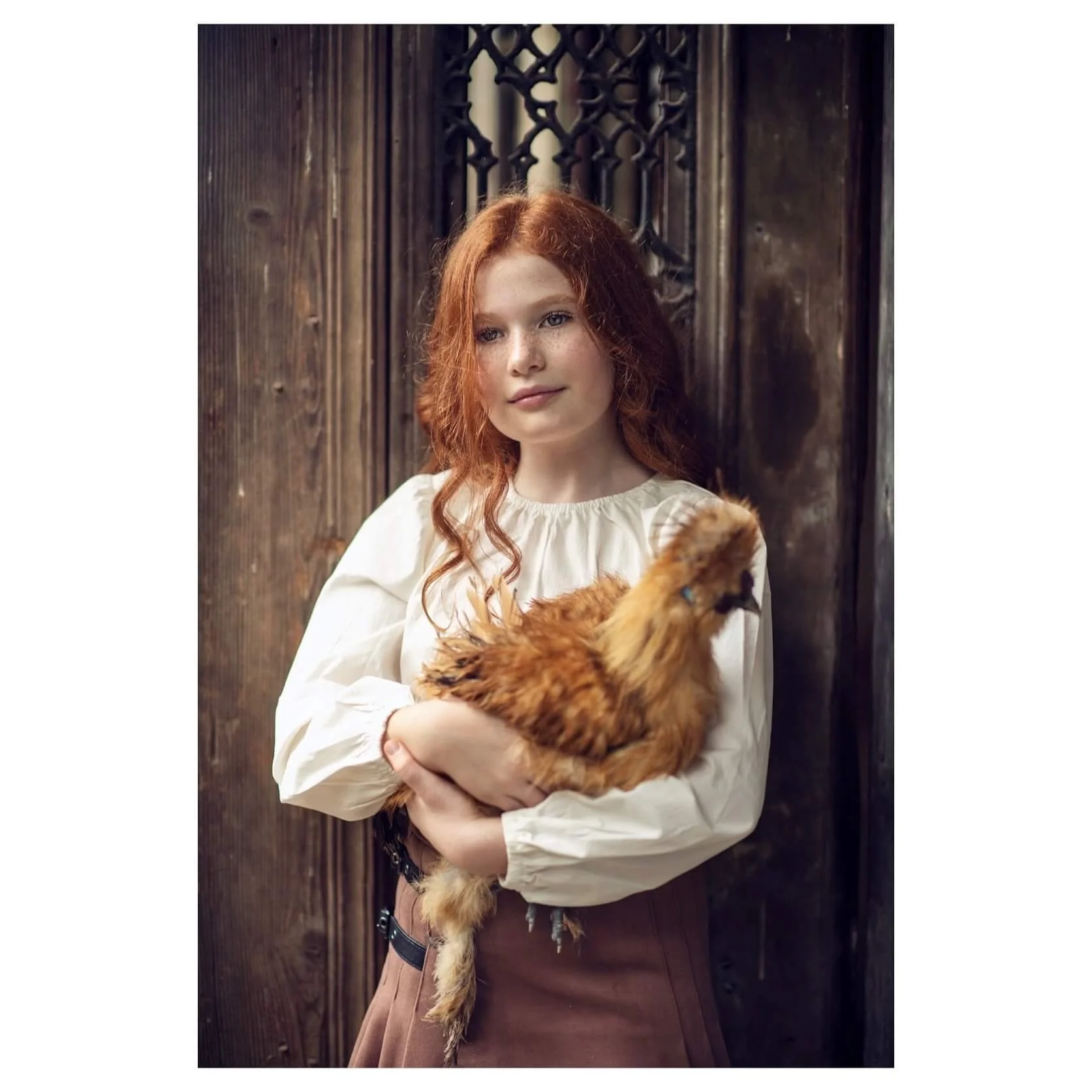 A young girl with red hair holding a chicken in front of a rustic wooden wall.
