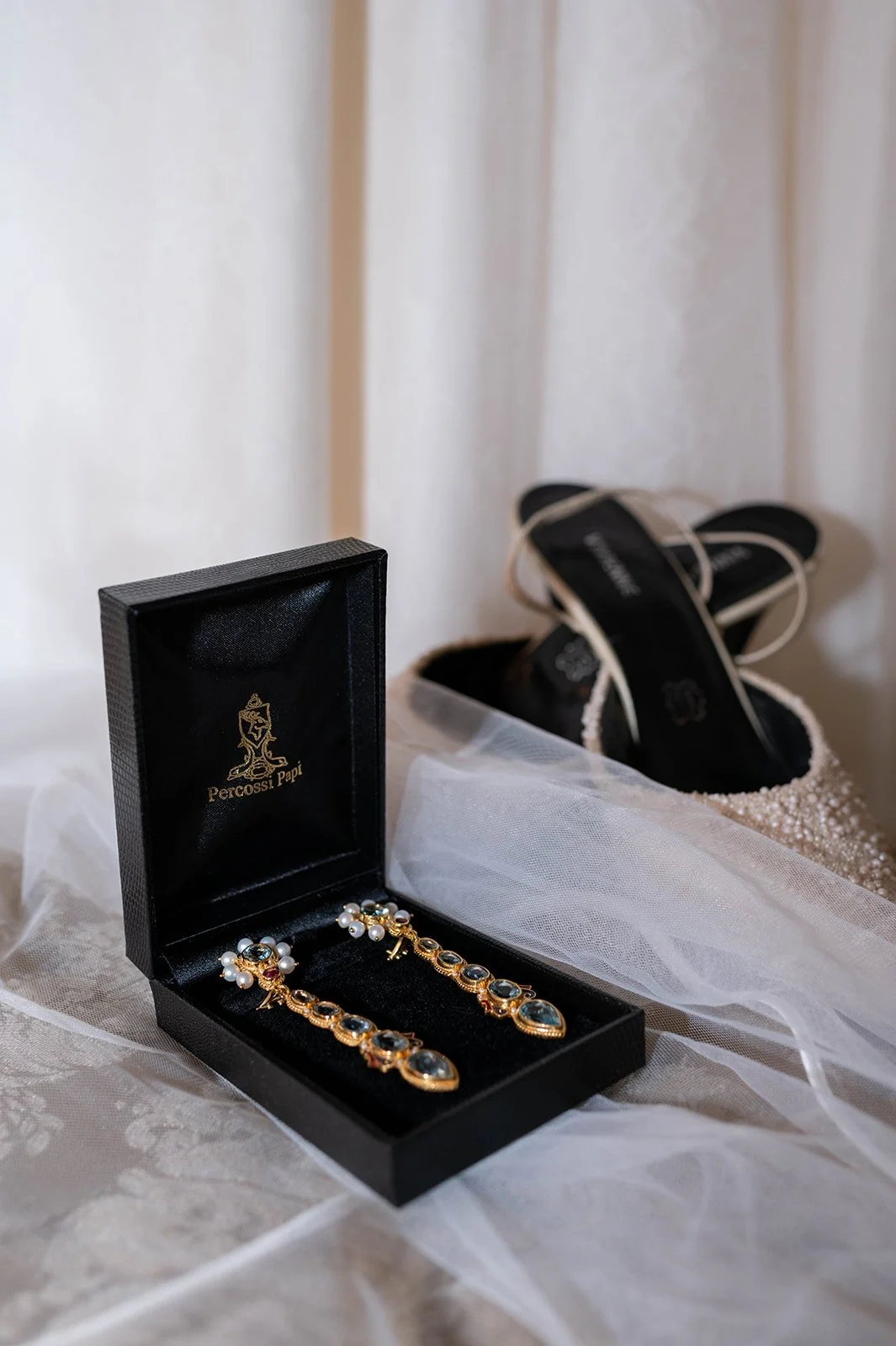 A pair of black and white high heel shoes, a black jewelry box with pearl and gold earrings, and a white veil on a textured fabric surface.