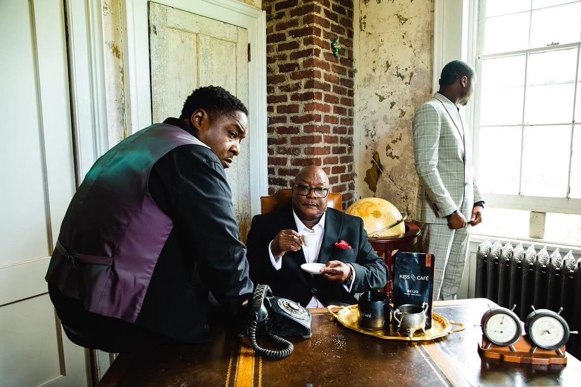 Three men in a vintage office setting with exposed brick walls. One man is sitting at a desk with a telephone and drinking from a small cup, another man is leaning over the desk talking to him, and the third man is standing near a window looking outs