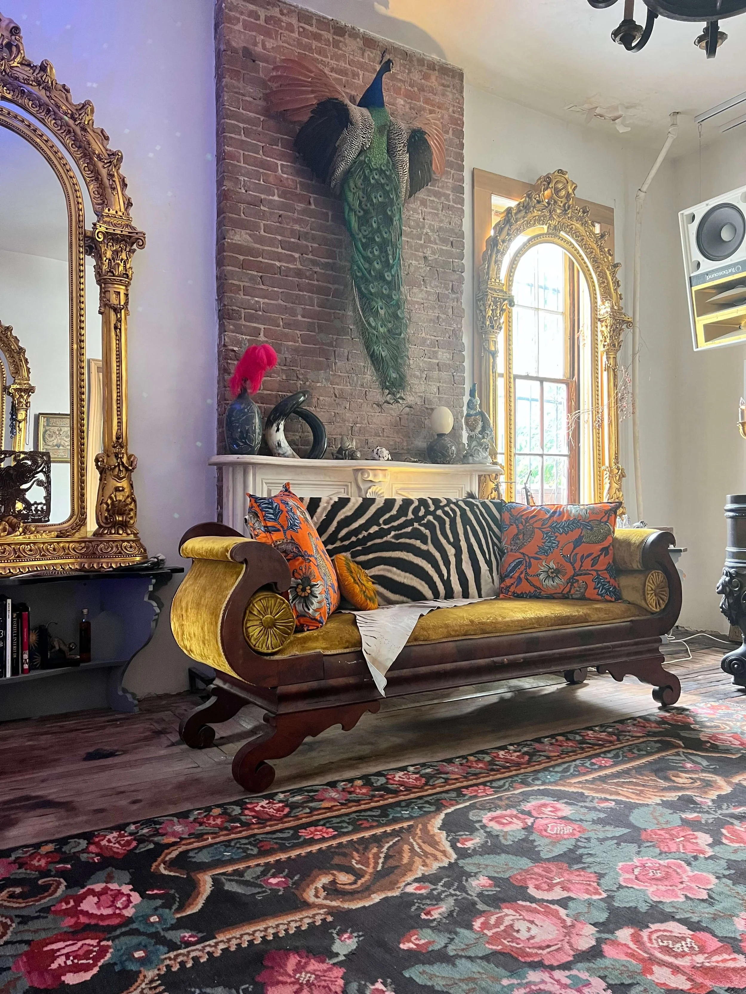 Living room decorated with vintage furniture, gold-framed mirrors, eclectic art, patterned throw pillows, and a peacock mounted on a brick wall.