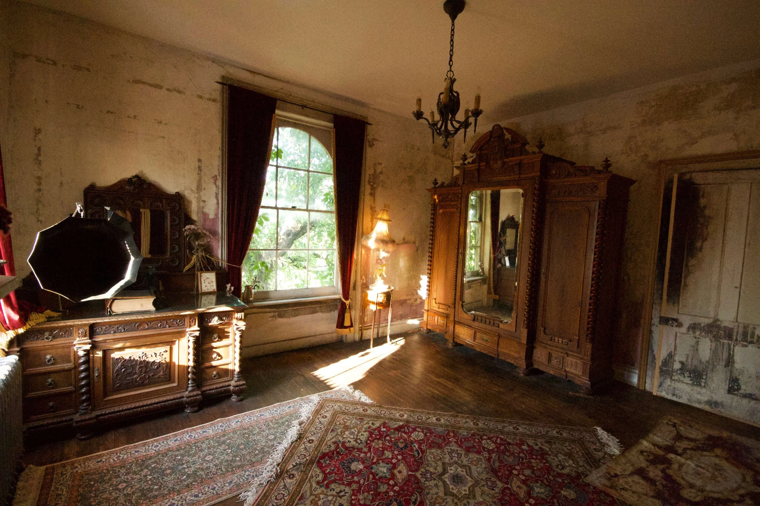 An old, vintage bedroom with antique wooden furniture, a large window with red curtains, a chandelier, worn walls, and oriental rugs on a wooden floor.