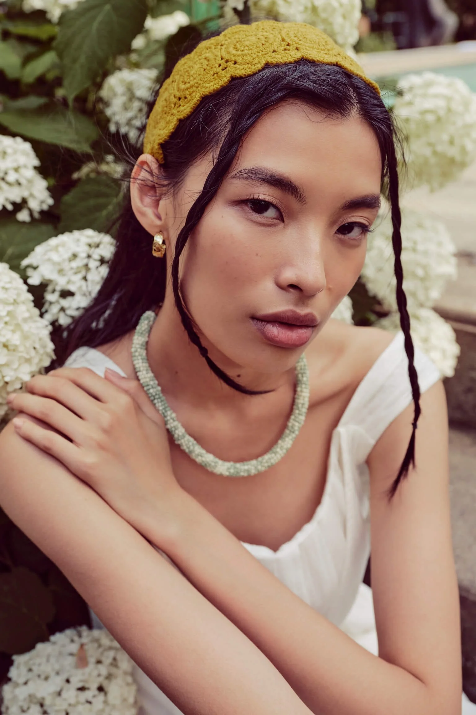 A young woman with black hair and wearing a yellow headband, white top, a beaded necklace, and earrings, posing among white flowers with an intense gaze.