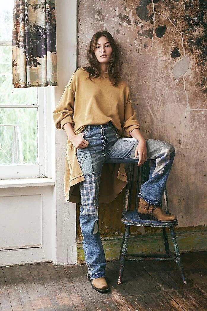 A young woman with dark brown hair and a beige oversized sweatshirt standing indoors next to a wall with peeling paint, with one foot on an old wooden chair. She is wearing cuffed patchwork jeans and tan ankle boots, with a neutral expression.
