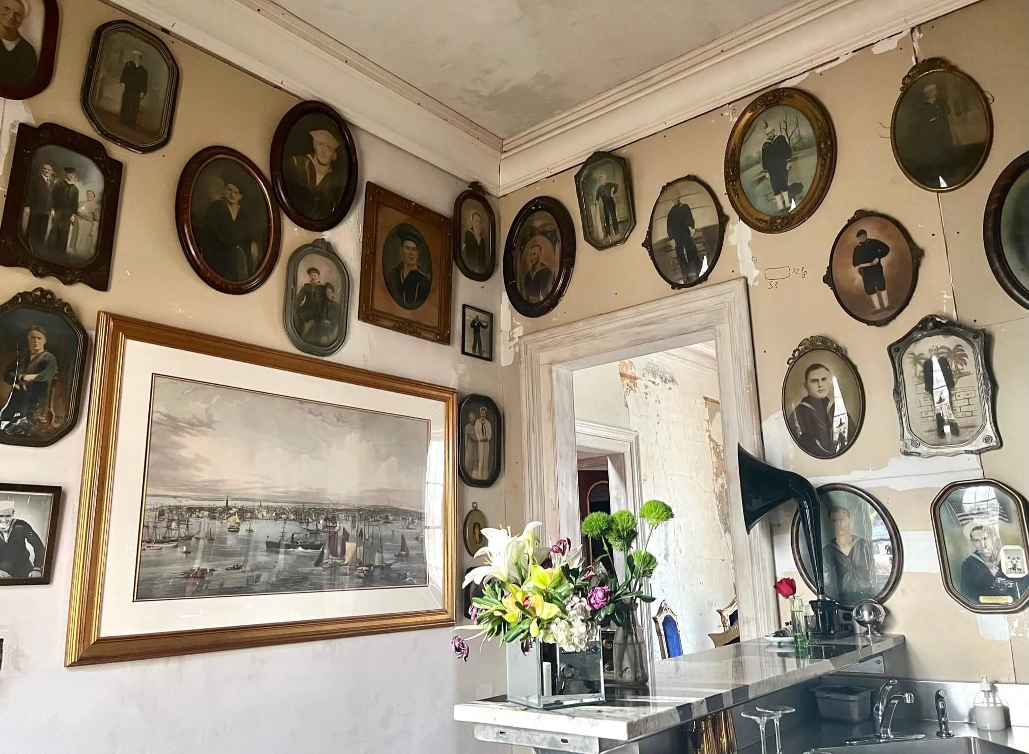 A wall decorated with numerous vintage oval picture frames containing black-and-white photographs of people in formal attire, a large framed painting of a harbor scene, a table with a vase of flowers and a gramophone, a doorway with peeling paint, an