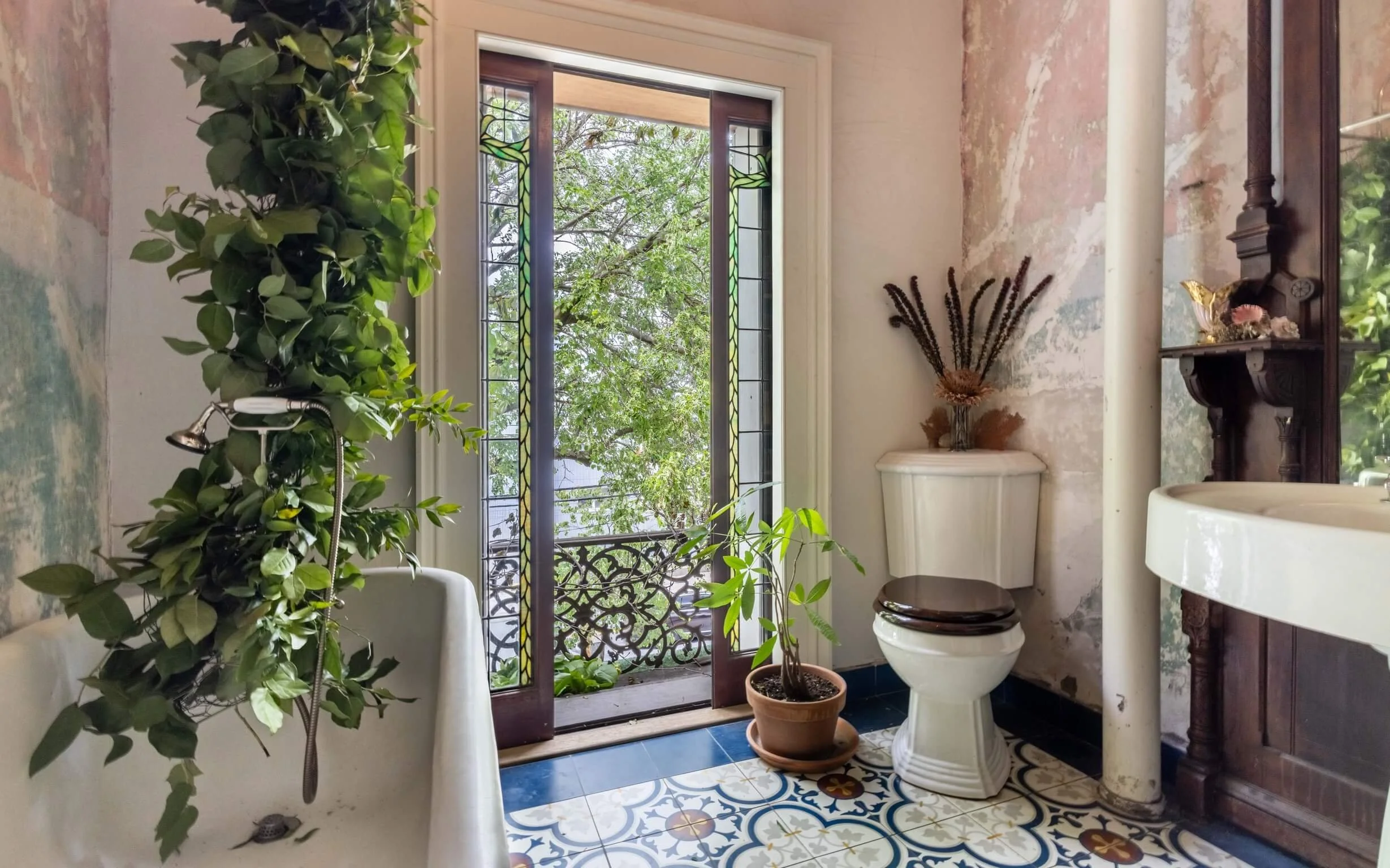 Bathroom with a window, potted plants, a white toilet with a dark wooden seat, a decorative tiled floor, and antique decor.